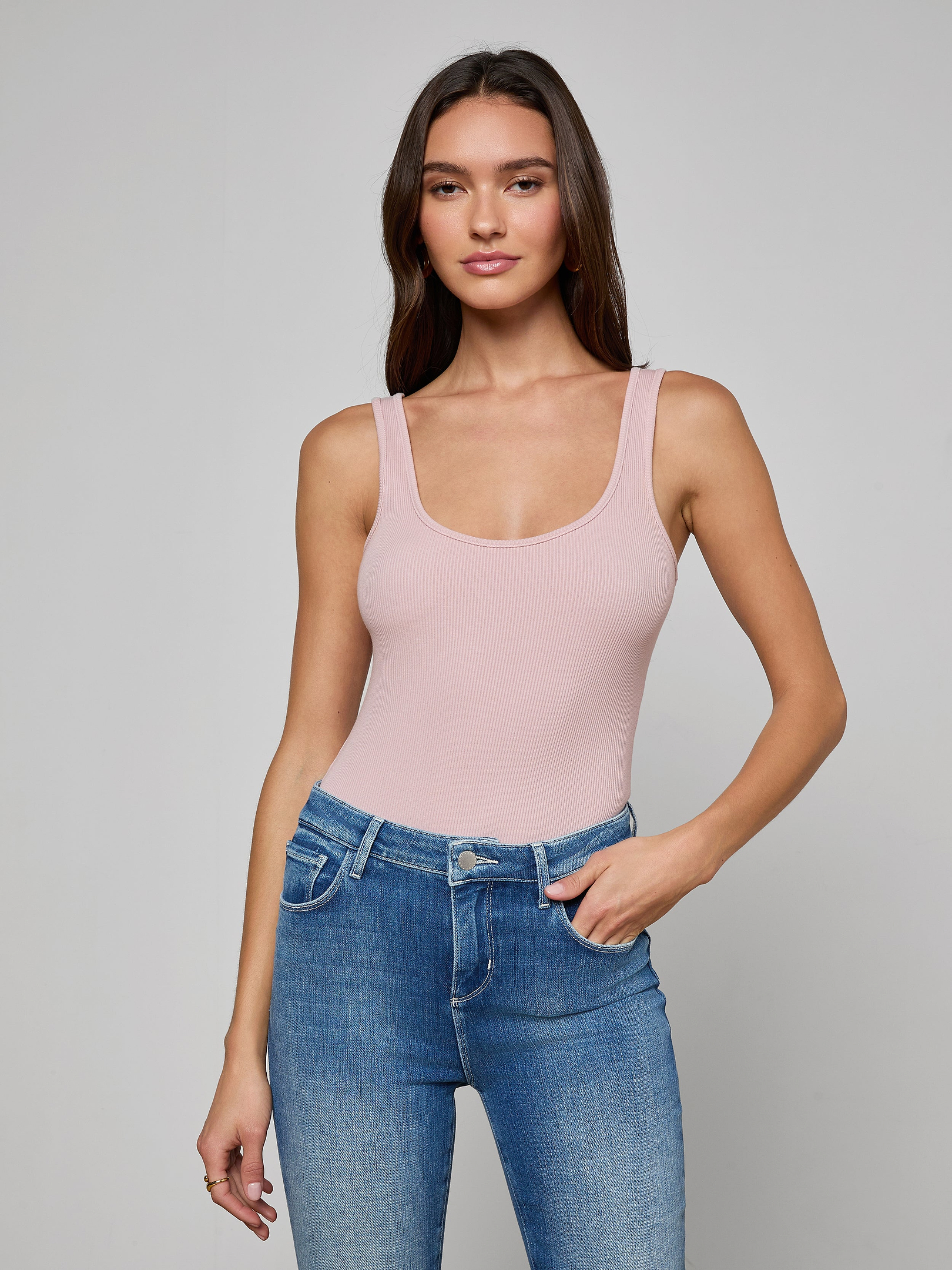 A woman with long brown hair stands against a plain light background, looking at the camera with a slight smile while wearing the Nica Scoopneck Bodysuit in light pink and blue jeans.