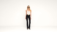 A woman wearing sunglasses, a white tank top, black pants, and black heels stands against a plain white background, facing forward with a neutral expression.