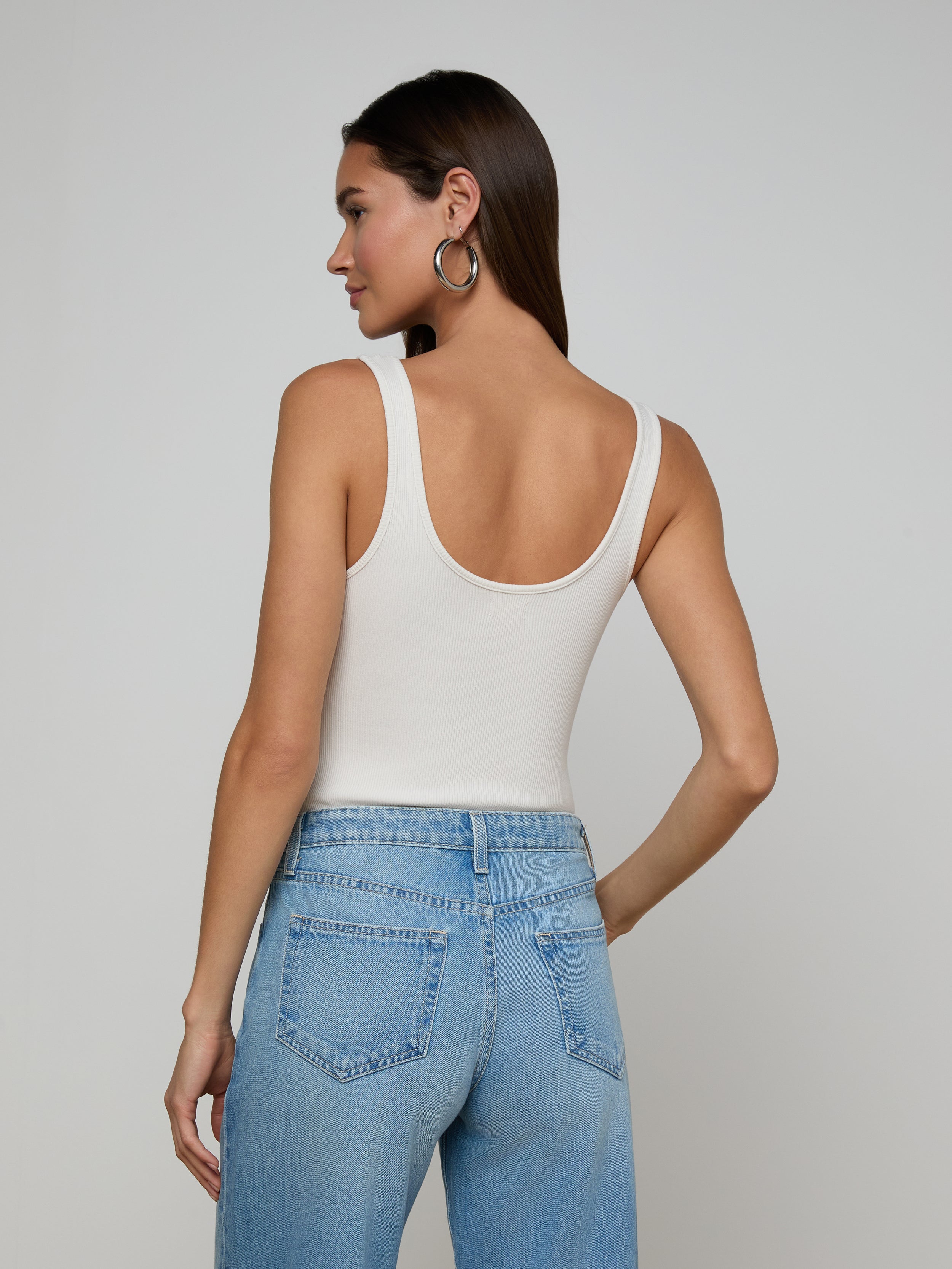 A woman with long blonde hair stands facing away, wearing a sleeveless beige Nica Scoopneck Bodysuit tucked into high-waisted black pants with a black textured belt, set against a plain light background.