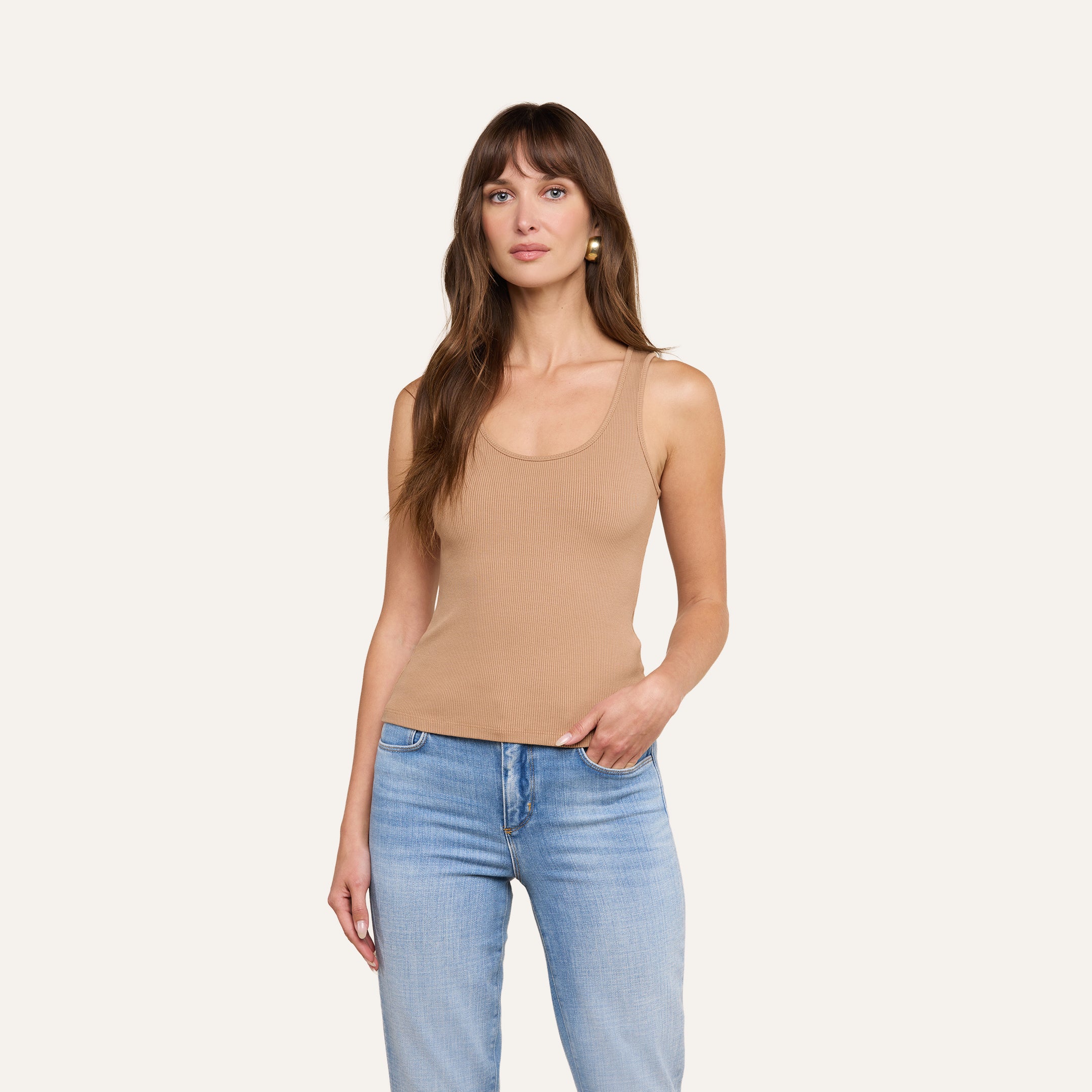 Une femme aux longs cheveux châtains porte le débardeur Adira, de couleur beige clair, avec un jean bleu clair, devant un fond beige uni, et regarde lobjectif avec une expression neutre.