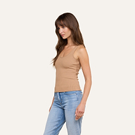 Une femme aux longs cheveux châtains se tient de profil, vêtue dun débardeur Adira à encolure dégagée et dun jean bleu clair, sur un fond beige uni.