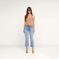 Une femme portant des lunettes de soleil, un débardeur Adira de couleur beige clair, un jean court bleu clair, une ceinture marron et des talons hauts beiges se tient debout devant un fond beige uni.