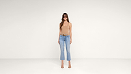Woman wearing sunglasses, a beige sleeveless top, blue cropped jeans, a brown belt, and heels stands against a plain white background.