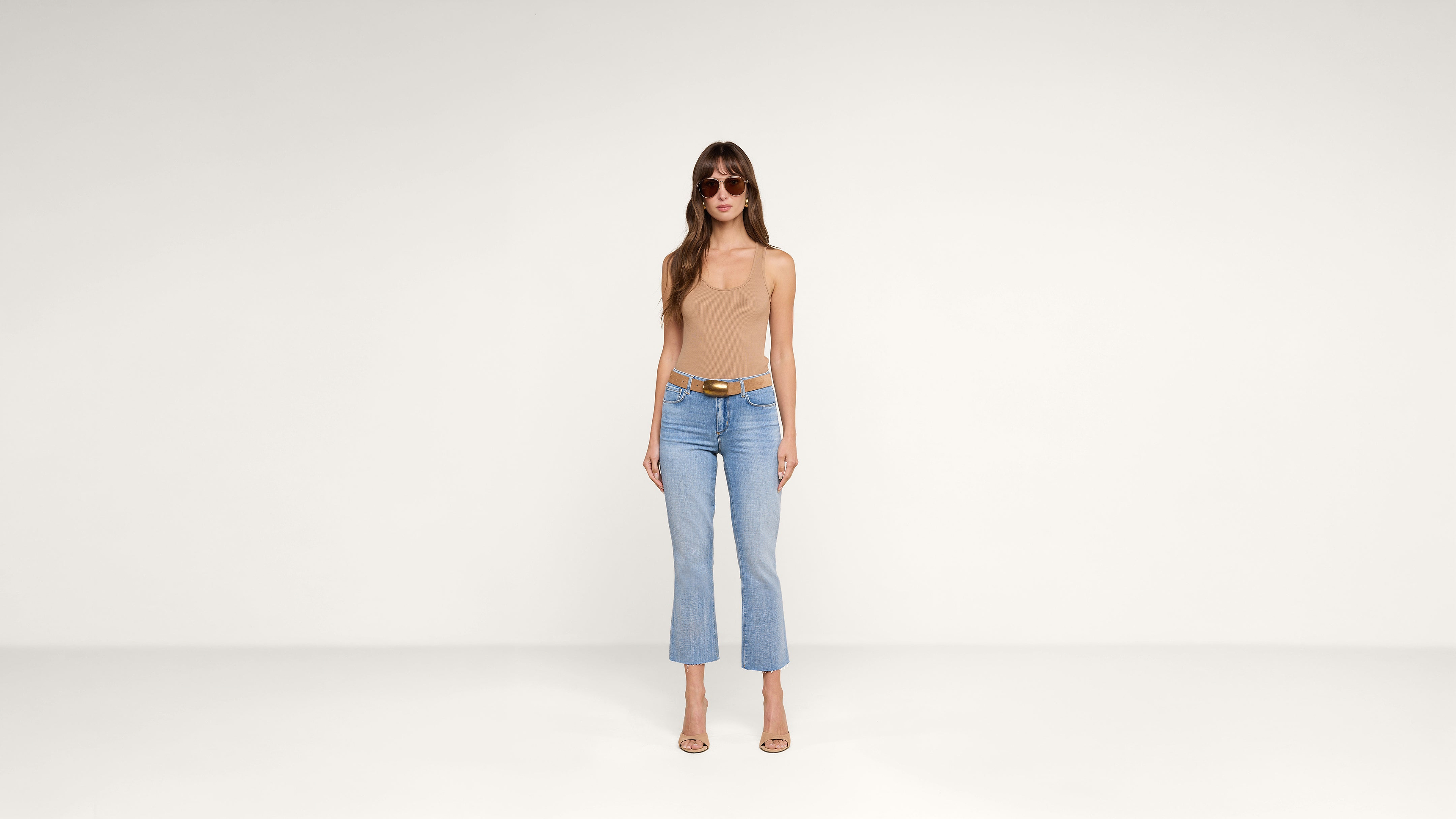 Woman wearing sunglasses, a beige sleeveless top, blue cropped jeans, a brown belt, and heels stands against a plain white background.