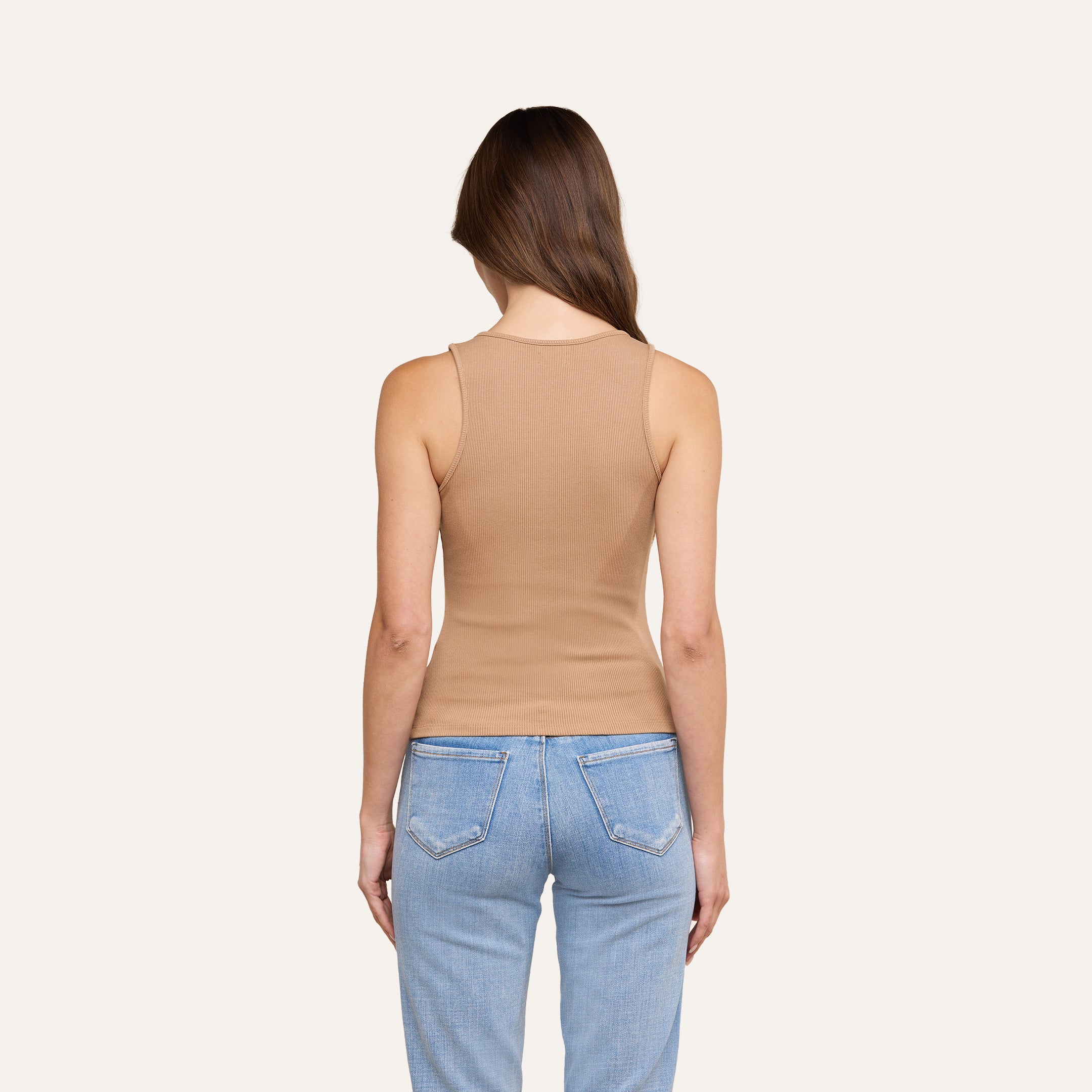 Une femme aux longs cheveux châtains, de dos, porte un débardeur Adira ajusté à côtes et un jean bleu clair, sur un fond beige uni.