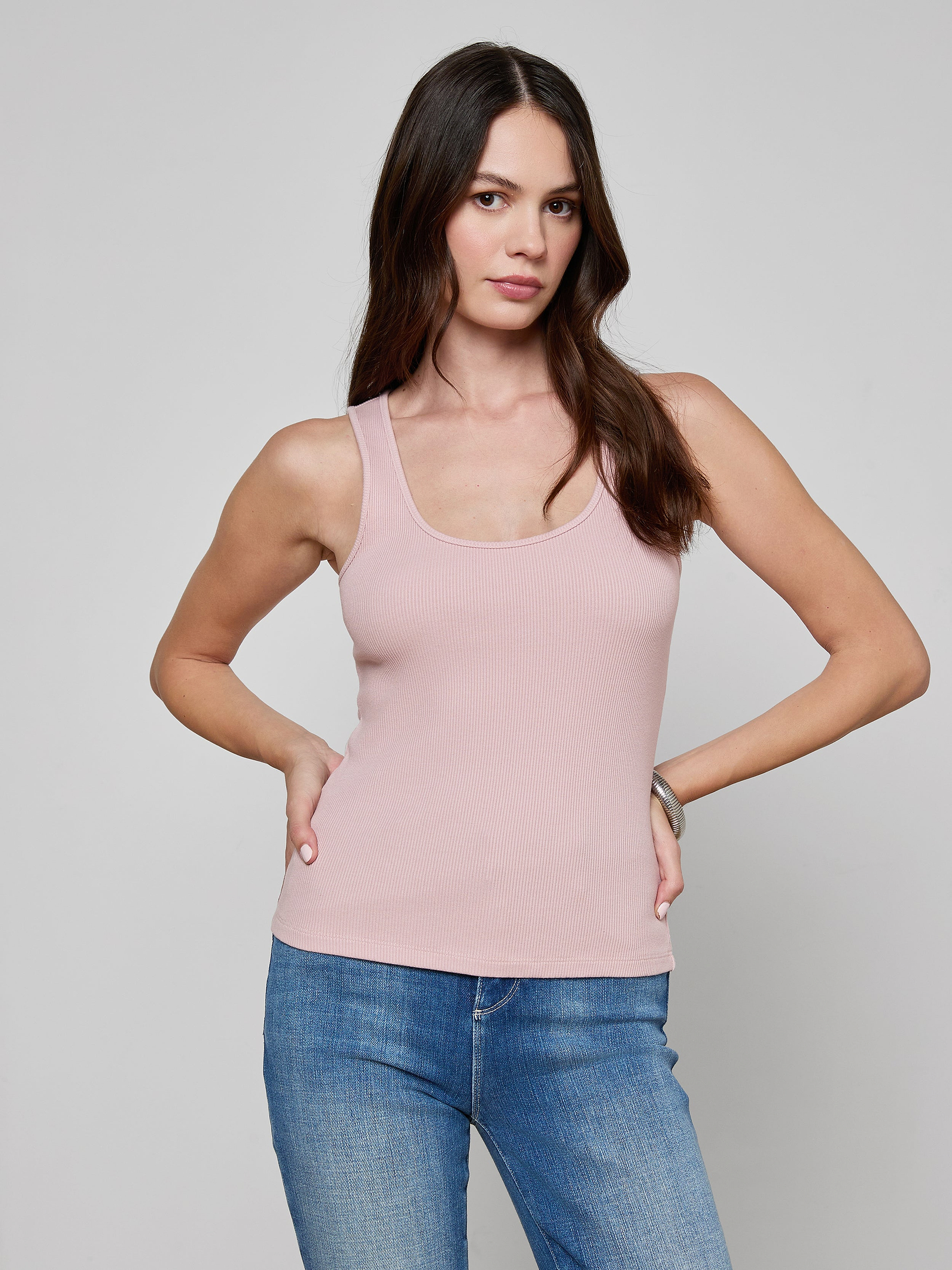 A woman with long brown hair wearing the Adira Scoopneck Tank in light pink and blue jeans stands with one hand on her hip, posing against a plain off-white background.