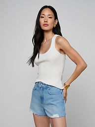 A woman stands against a plain background, wearing the Adira Scoopneck Tank in white, beige pants with a belt, and gold earrings. She has long blonde hair and a neutral expression, facing slightly to the left.