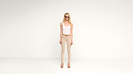 A woman wearing sunglasses, a white tank top, beige pants, and beige heels stands against a plain white background.