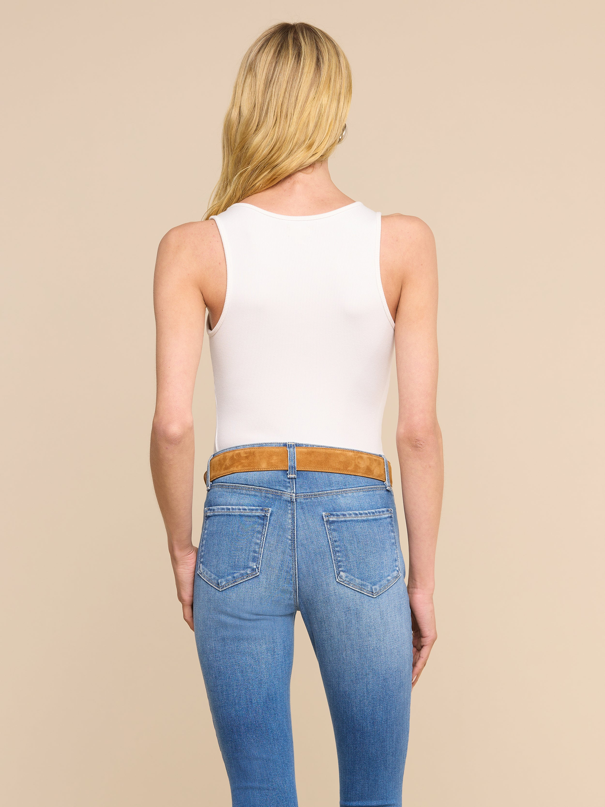 A woman with long blonde hair, wearing the Adira Scoopneck Tank in white and blue jeans with a brown belt, stands facing away from the camera against a plain light background.