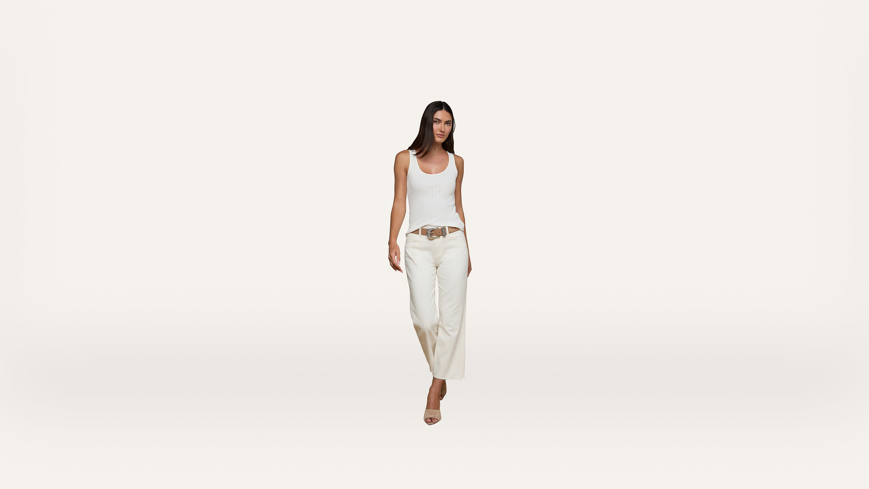 A woman with long dark hair wearing a white tank top, wide-leg white pants, a brown belt, and beige heels walking forward against a plain white background.