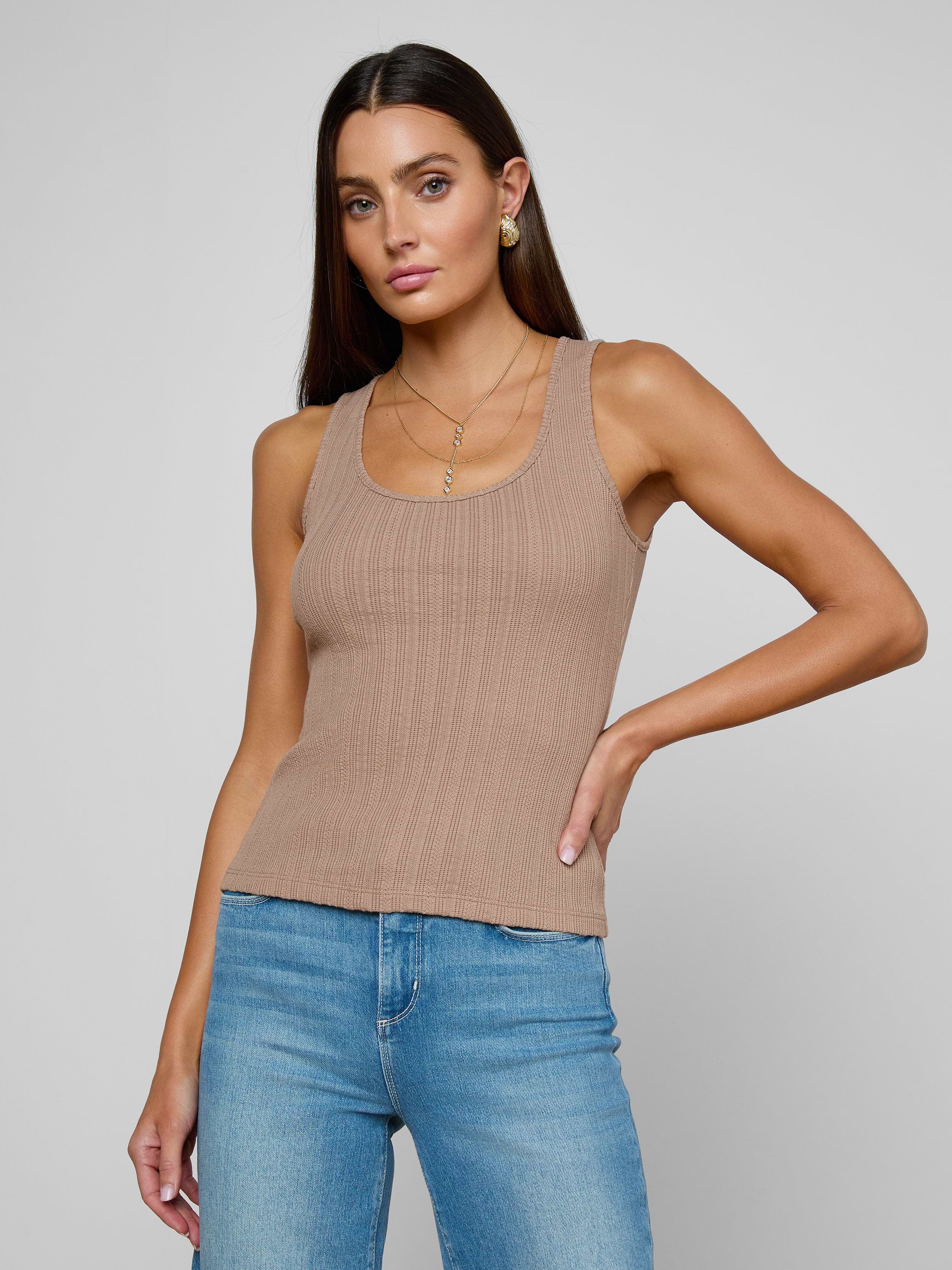 A woman wearing the Adira Scoopneck Tank—a fitted, ribbed beige organic cotton top—paired with blue jeans stands against a plain light background, one hand on her hip and a neutral expression.