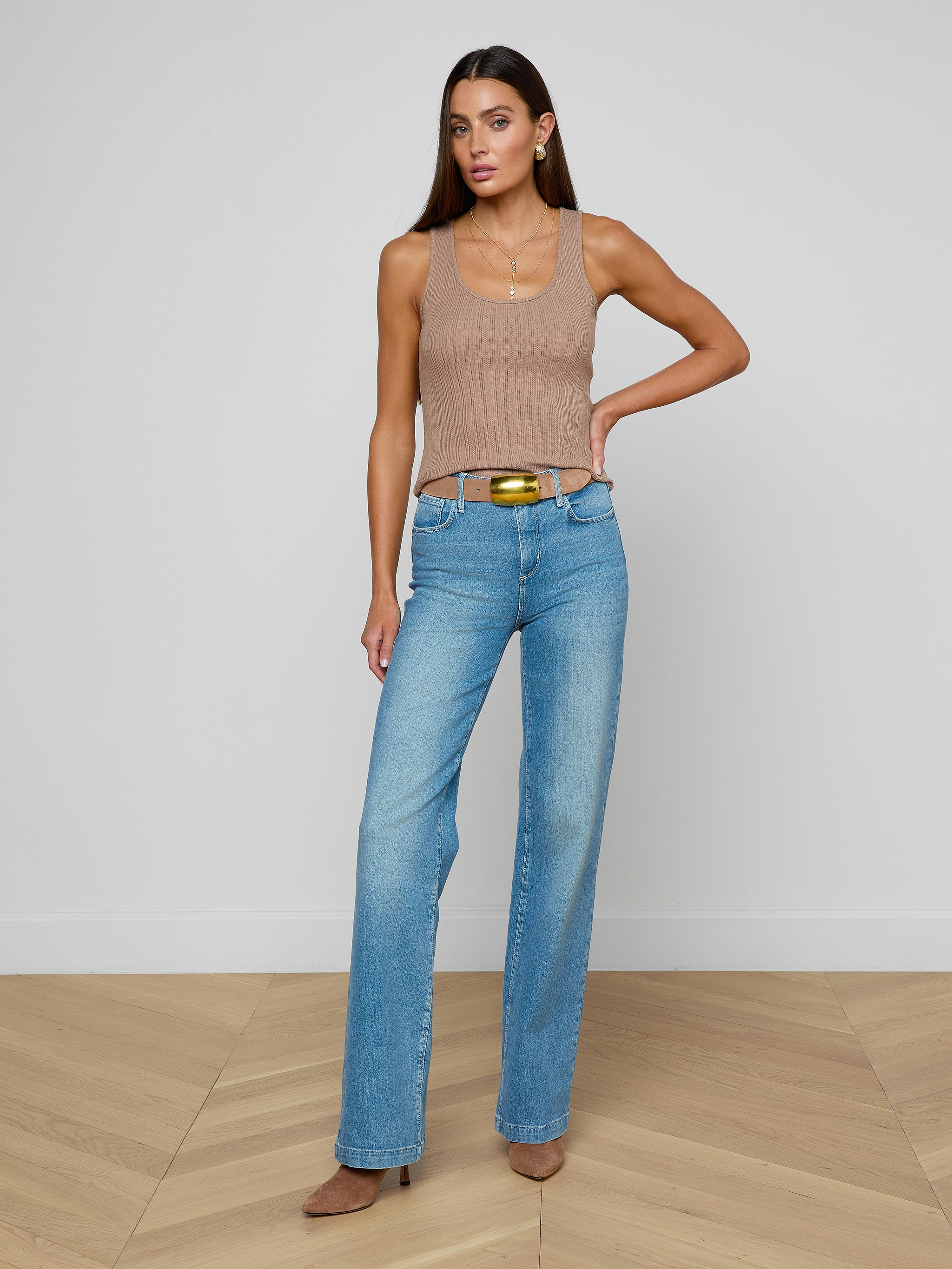 A woman models the Adira Scoopneck Tank— a fitted, sleeveless beige top with a scoop neckline— paired with blue jeans and brown heels, standing with one hand on her hip against a plain white background.