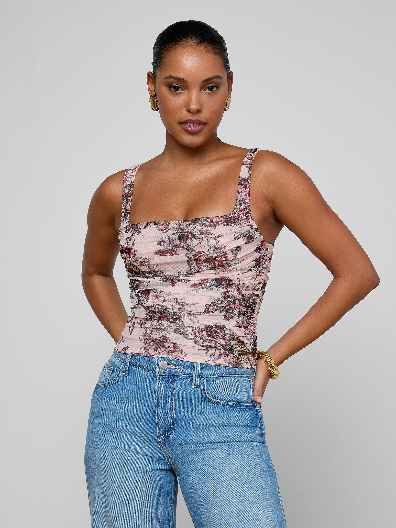 A woman wearing the Rasha Shirred Mesh Tank, featuring a pink floral pattern and square neckline, pairs it with blue jeans as she stands facing forward with her hands in her pockets against a plain light background.