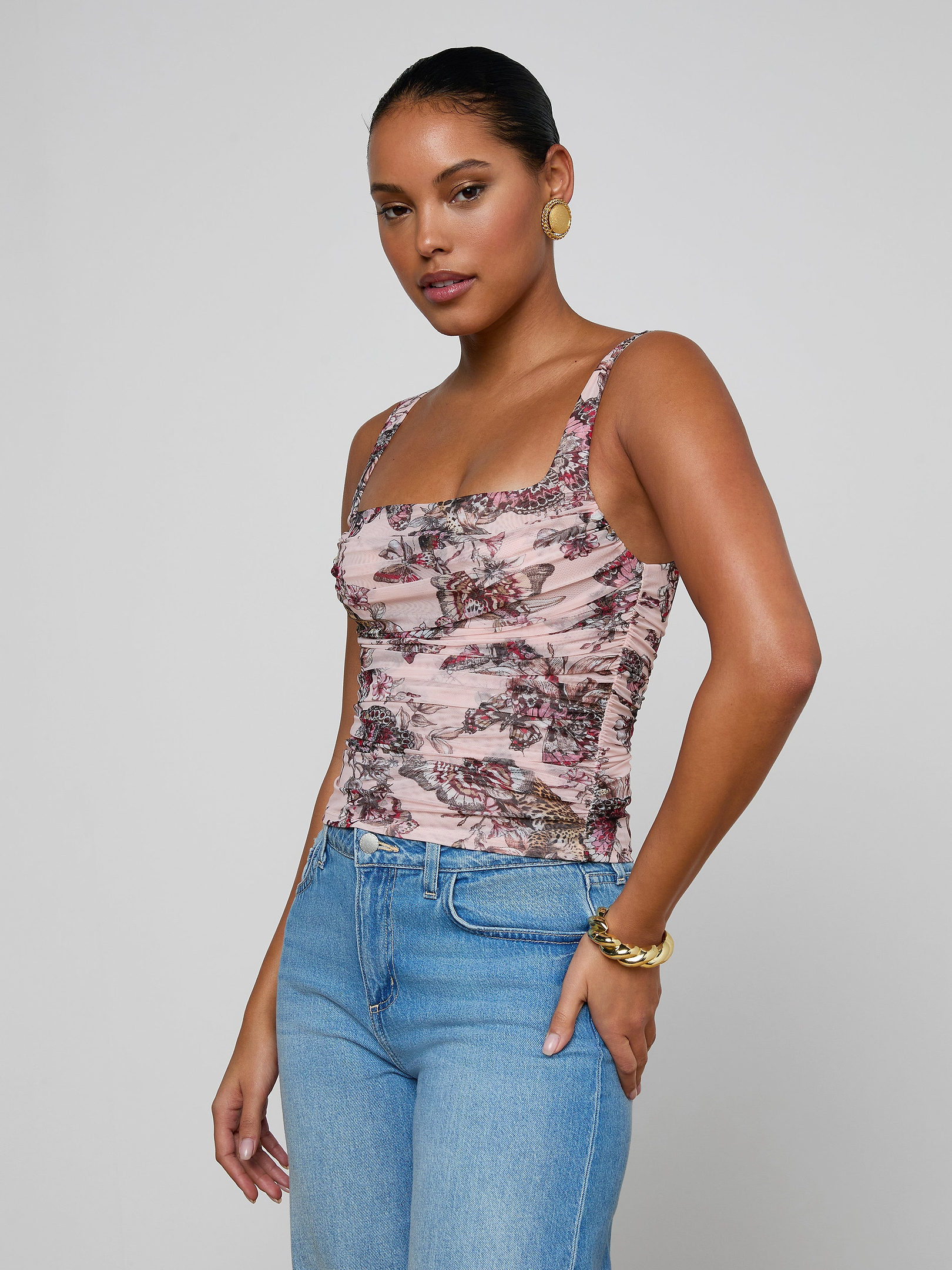 A woman wearing the Rasha Shirred Mesh Tank, which features a square neckline and a butterfly-jungle print, paired with blue jeans, stands with one hand in her pocket against a plain light background.