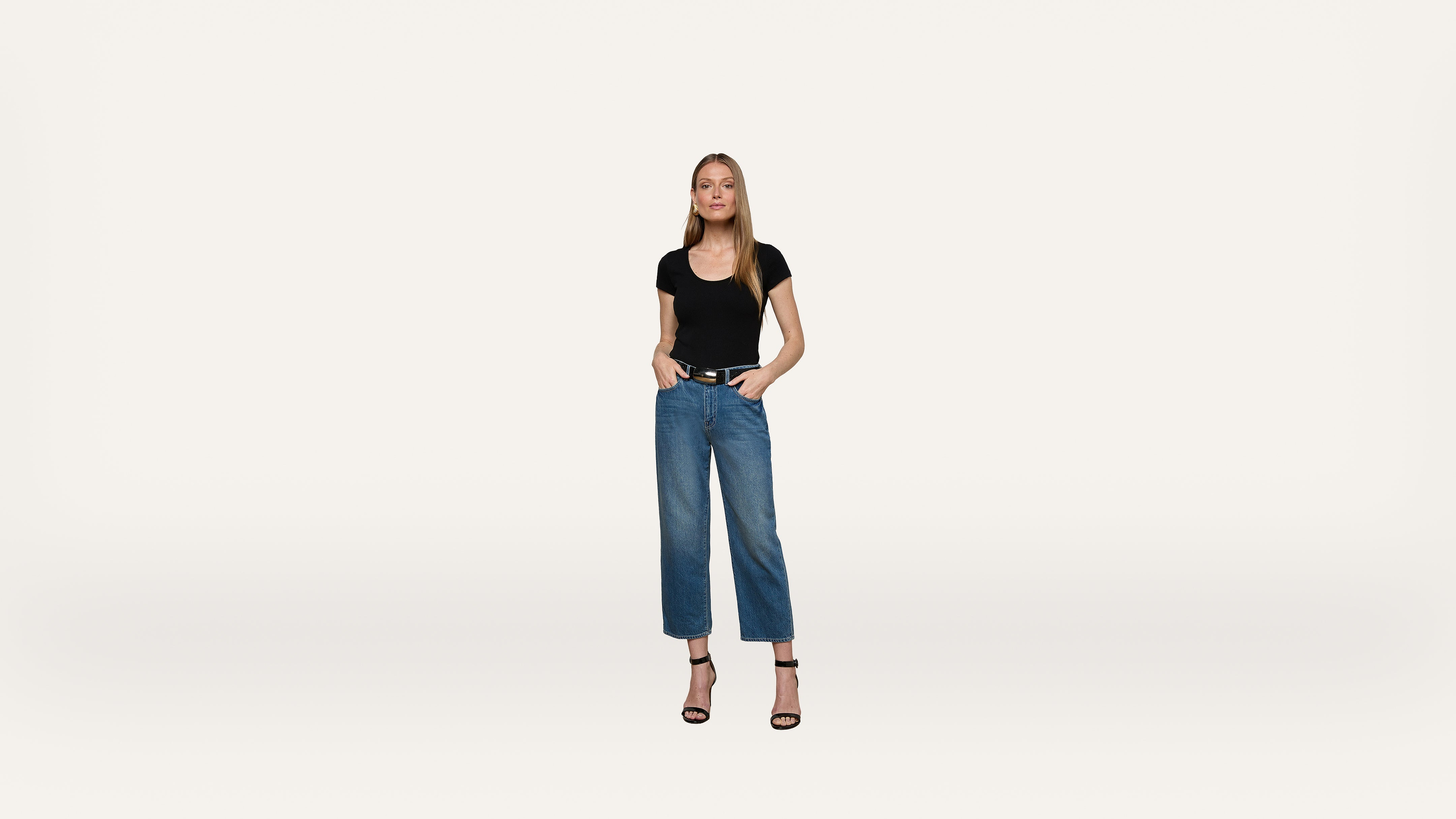 A woman stands against a plain white background, wearing a black t-shirt, blue jeans, and black high-heeled sandals. Her hands are in her pockets, and she faces forward with a neutral expression.
