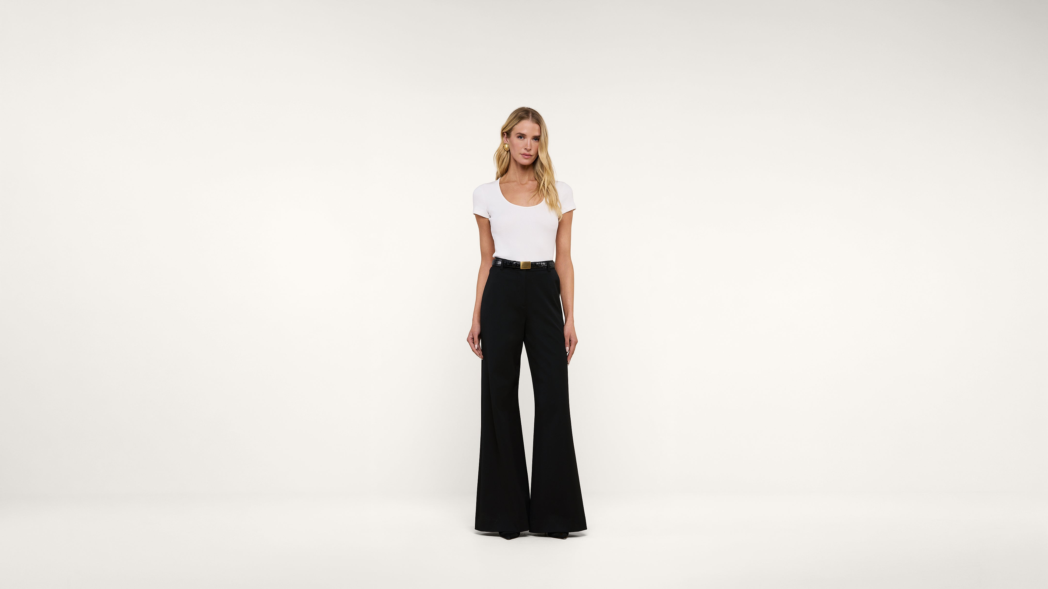 Woman standing against a plain white background, wearing a white short-sleeve top tucked into high-waisted, wide-leg black pants with a black belt. She has long, blonde hair and a neutral expression.