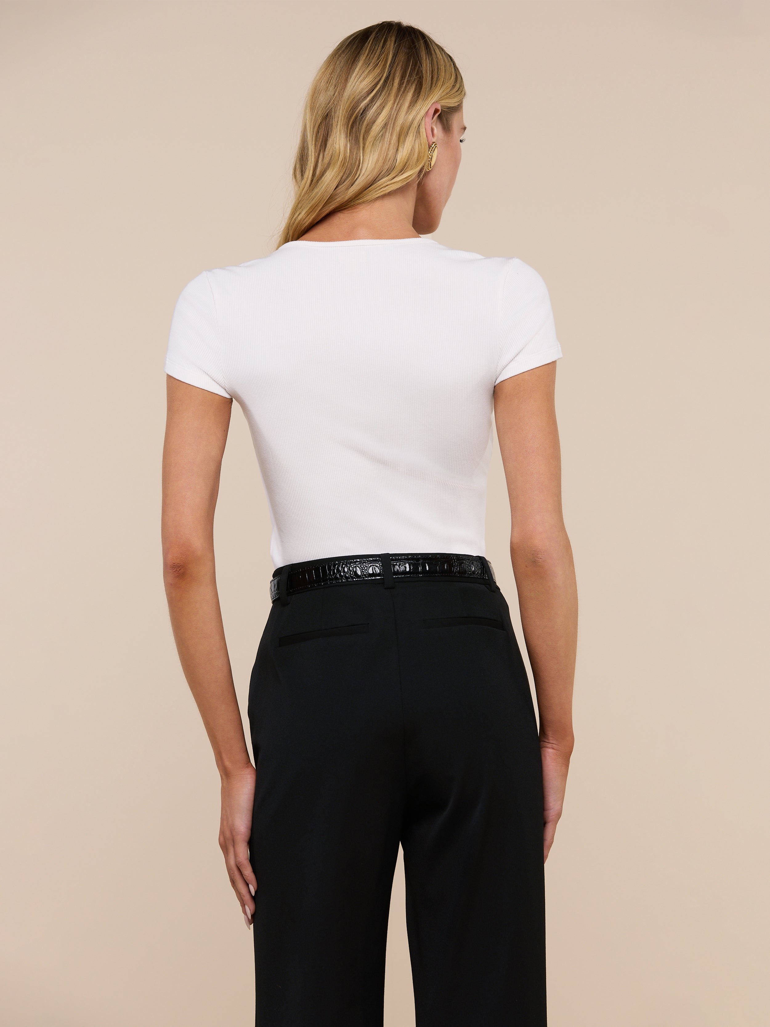 A woman with long blonde hair is shown from the back, wearing the fitted Brynlee Scoopneck Tee in white with short sleeves and high-waisted black pants with a belt.