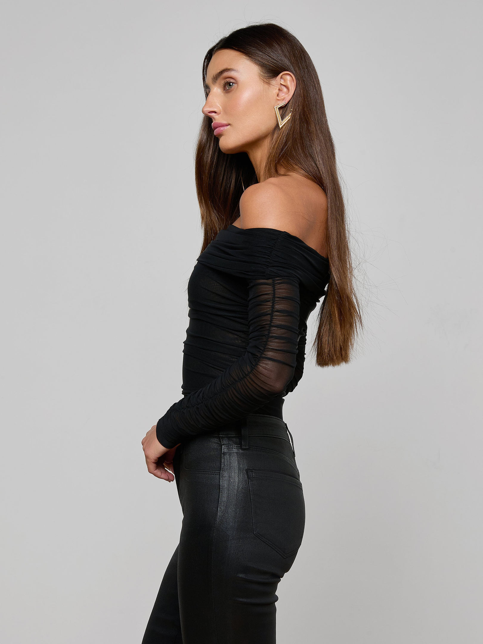 A woman in profile wearing the Poppie Off-The-Shoulder Bodysuit—with stretch mesh sleeves—paired with black fitted pants, standing against a plain, light background.