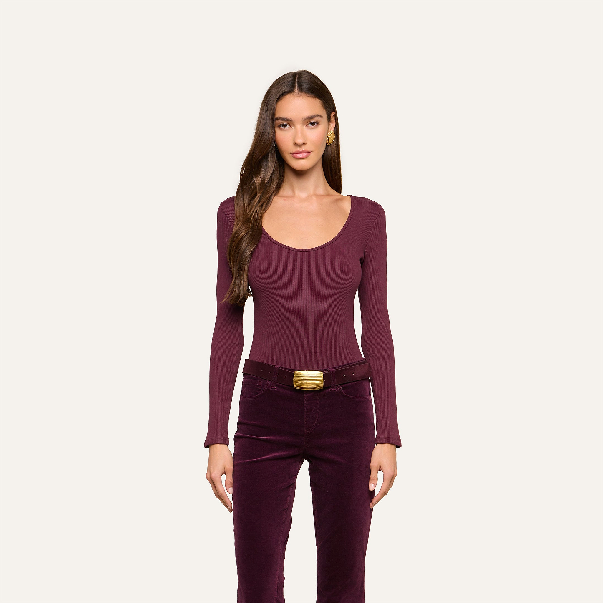Woman with long brown hair, wearing the Elara Scoopneck Bodysuit—a fitted long-sleeve burgundy top crafted from stretch cotton modal—and high-waisted burgundy pants with a large rectangular belt, stands facing forward against a plain light background.