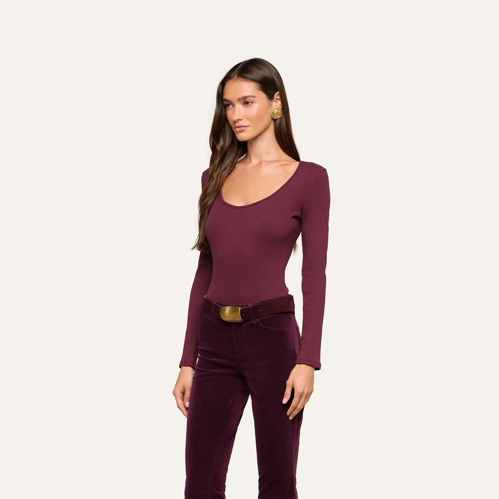 A woman with long brown hair is wearing the fitted Elara Scoopneck Bodysuit in burgundy, paired with matching velvet pants. She accessorizes with gold earrings and a belt, standing against a plain light backdrop. The body-contouring fit of the bodysuit beautifully enhances her silhouette.