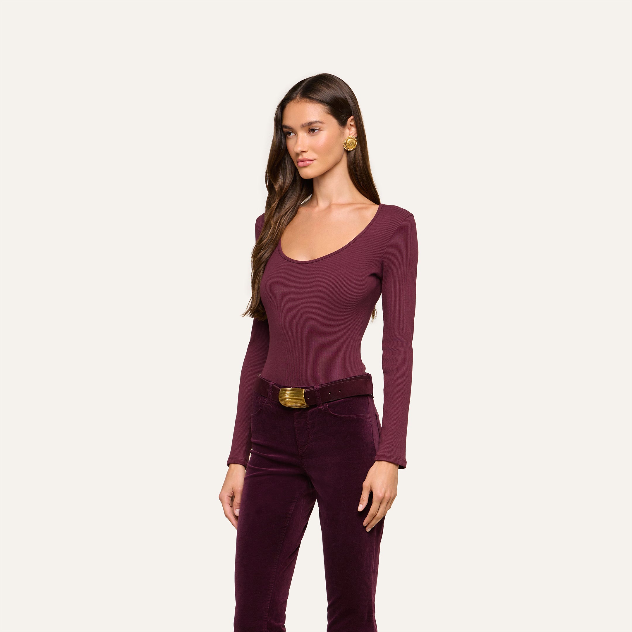 A woman with long brown hair is wearing the fitted Elara Scoopneck Bodysuit in burgundy, paired with matching velvet pants. She accessorizes with gold earrings and a belt, standing against a plain light backdrop. The body-contouring fit of the bodysuit beautifully enhances her silhouette.