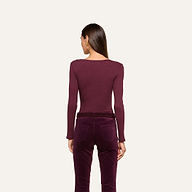 A woman stands with her back to the camera, wearing the Elara Scoopneck Bodysuit in maroon—crafted from stretch cotton modal—and high-waisted velvet pants in a coordinating shade, set against a simple light background. The look emphasizes the Elara’s sleek, body-contouring fit.