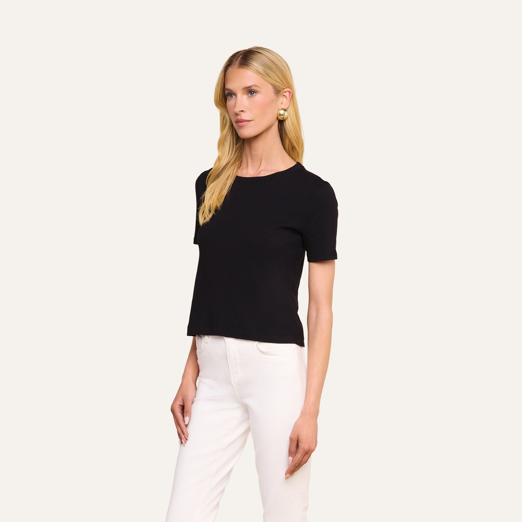Una mujer con el pelo largo y rubio lleva la camiseta corta de algodón «Stormy» en color negro, de corte semiholgado, combinada con unos pantalones blancos y unos pendientes dorados. Aparece de pie frente a un fondo liso de color beige, mirando ligeramente hacia la izquierda.