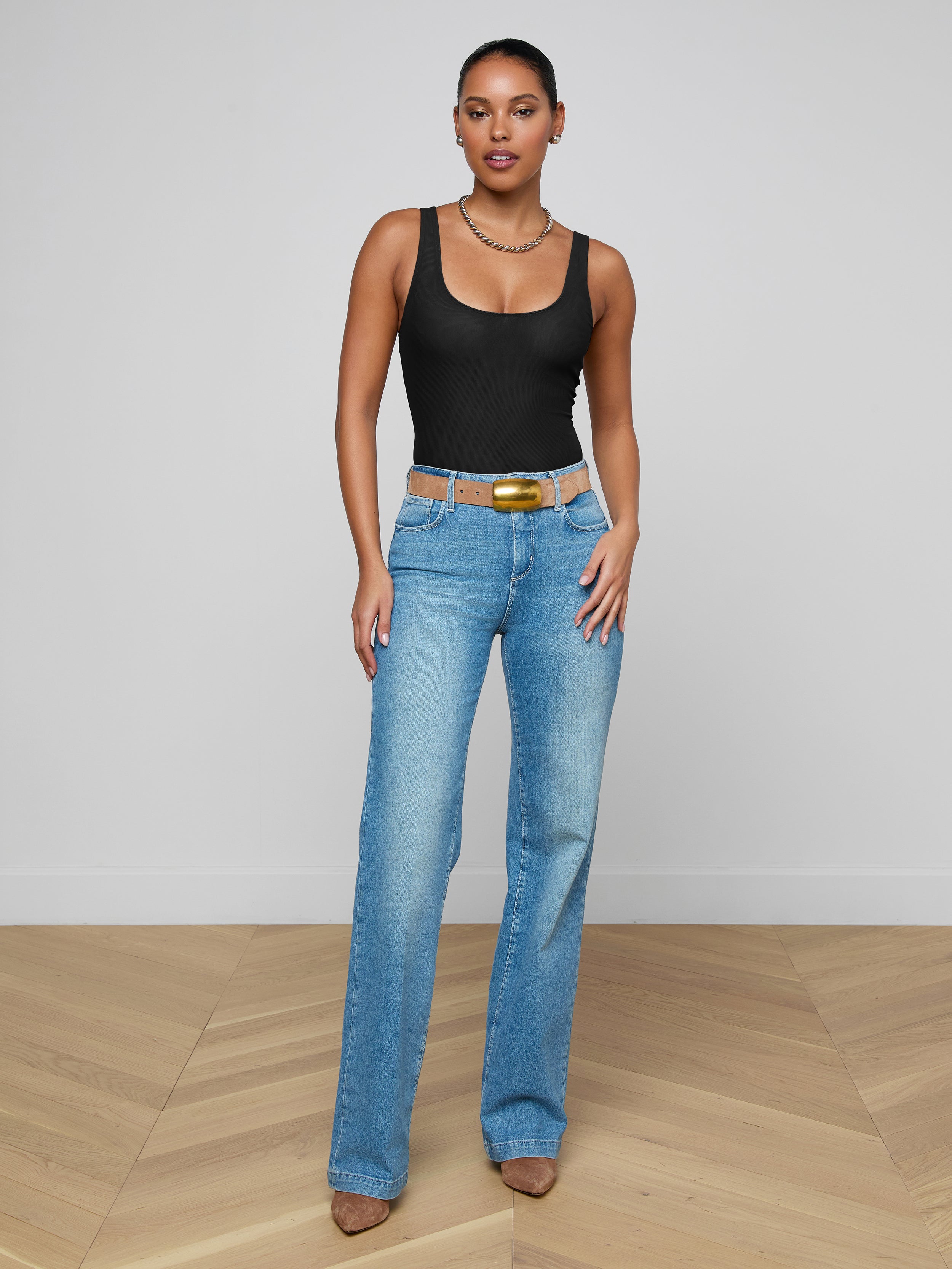 A woman wearing the Kalea Mesh Bodysuit, paired with blue high-waisted jeans and brown heels, stands against a plain white background.