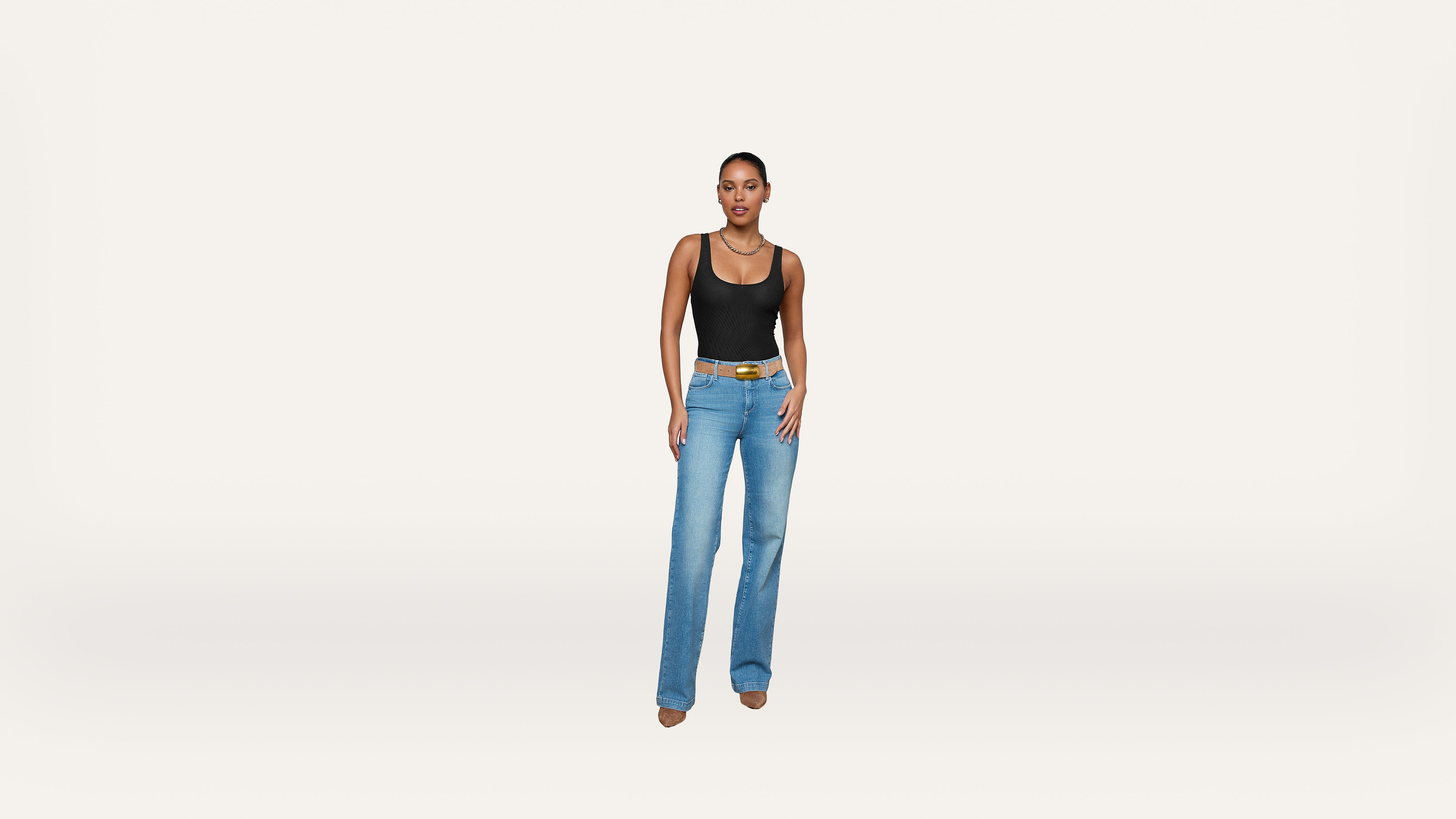 A woman stands against a plain white background, wearing a black tank top, blue jeans, a brown belt, and beige shoes.