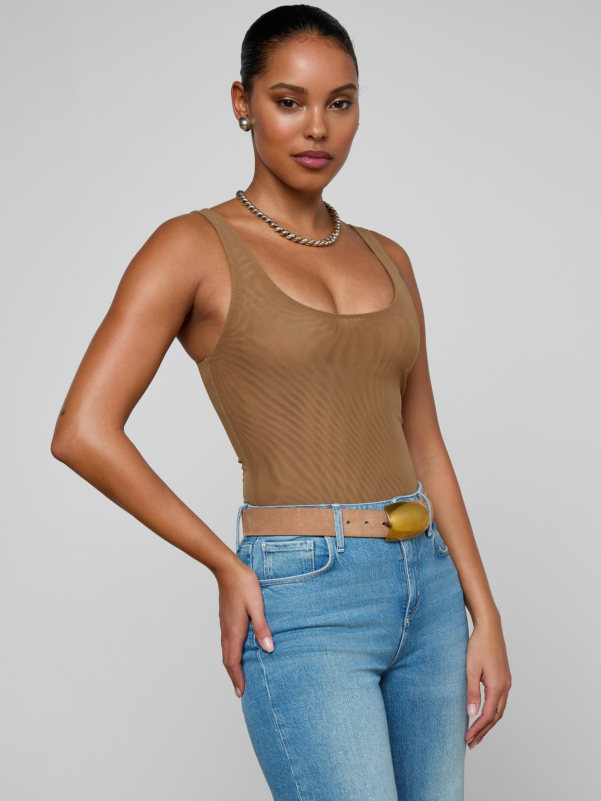 A woman with long brown hair is wearing the Kalea Mesh Bodysuit in a lightweight tan fabric, paired with blue jeans and a belt. She stands in profile against a plain light background, accessorized with gold earrings and sporting a neutral expression.