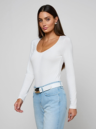 Woman with long brown hair wearing the Cassandra Ruched Bodysuit—a white long sleeve top with a scoop neckline—paired with light blue jeans and a white belt, standing against a plain light background.