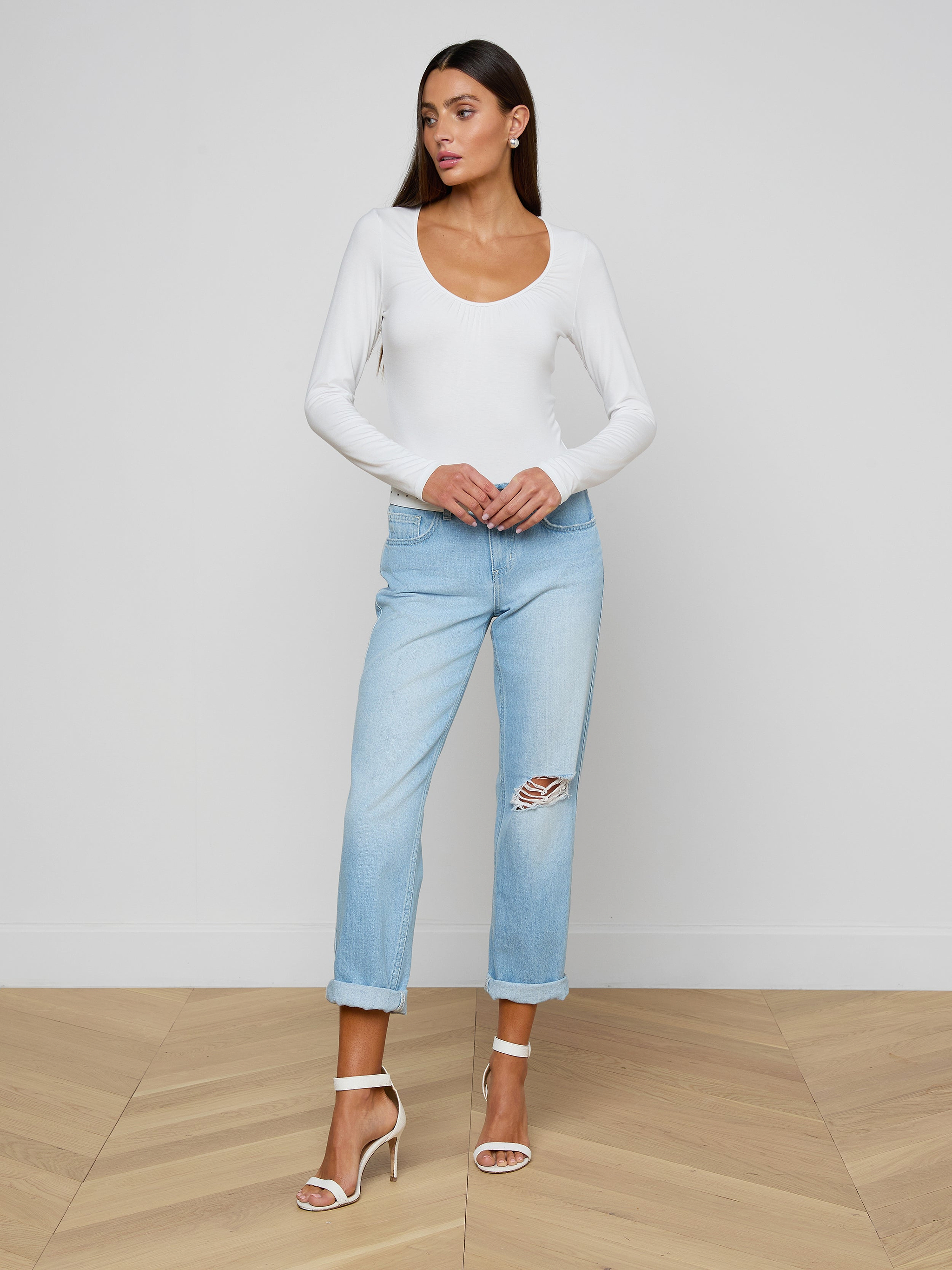 The woman is wearing the Cassandra Ruched Bodysuit, a white long-sleeve top with a scoop neckline, paired with light blue ripped jeans and high-heeled sandals. She stands against a plain light background, looking to the side with her hands resting on her jeans.