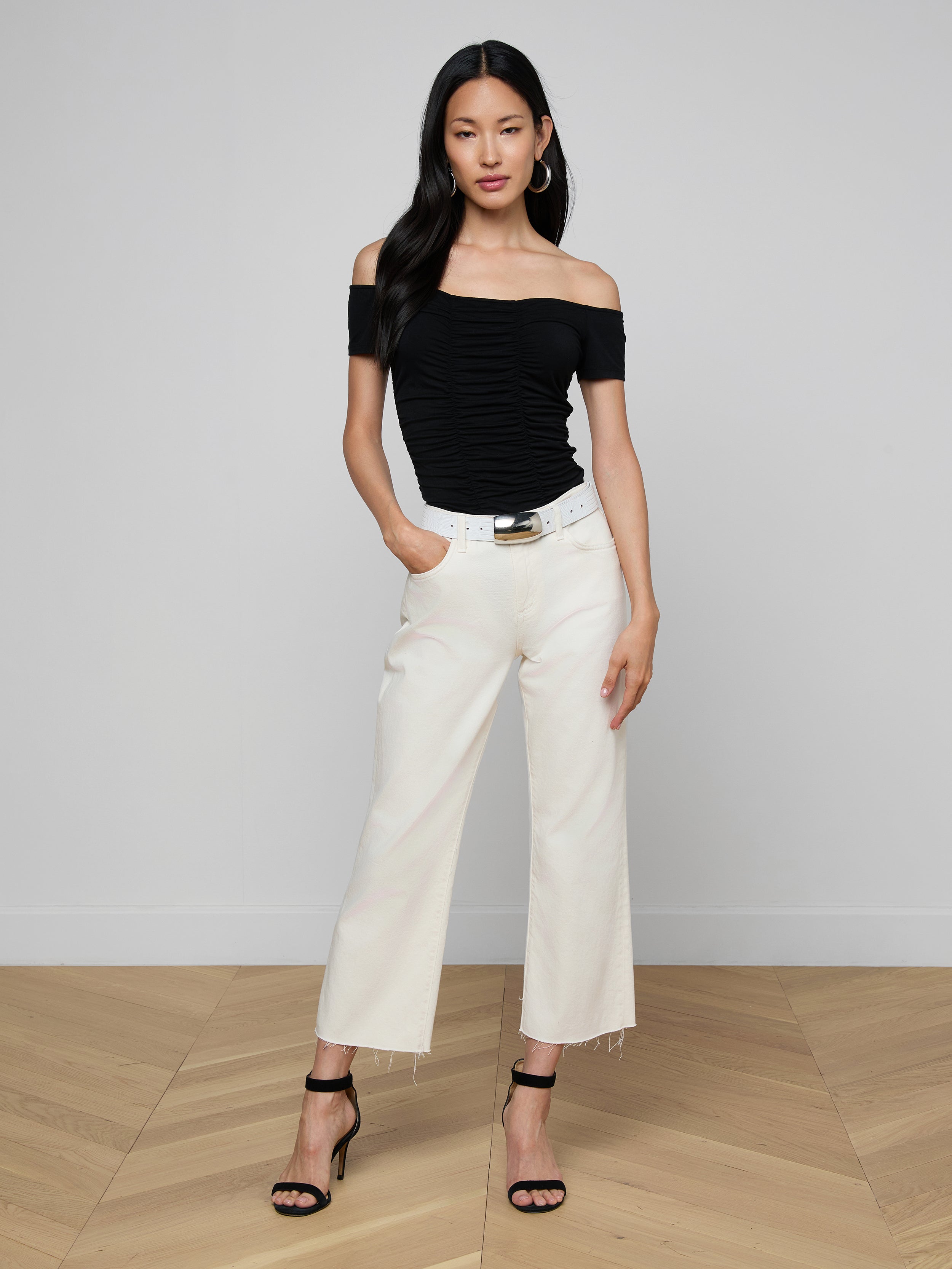 Woman wearing the Baliegh Ruched Bodysuit with an off-the-shoulder neckline, paired with white wide-leg pants, a black belt, and black high-heeled sandals, standing against a plain light background with her hands in her pockets.