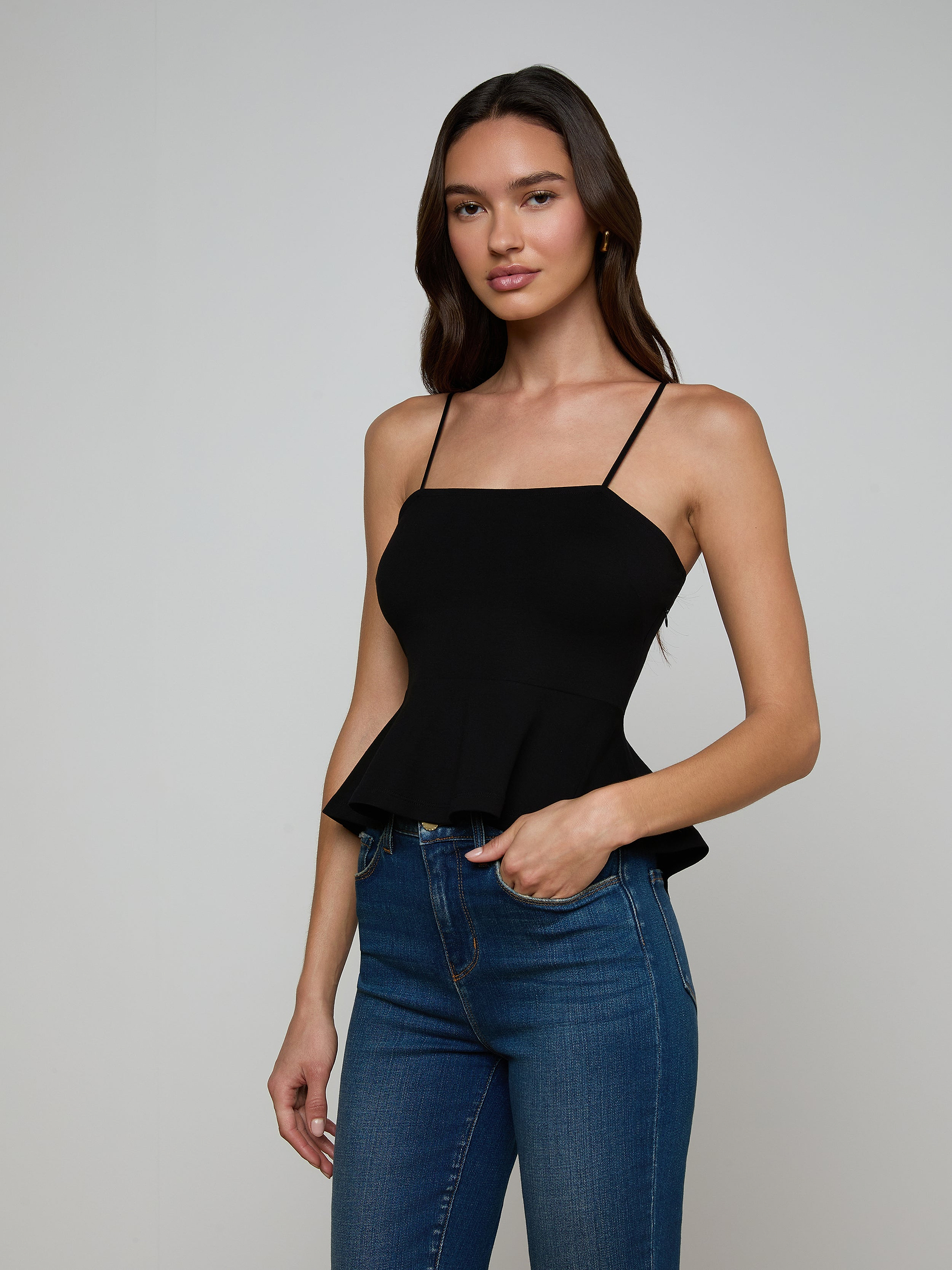 A woman wearing the Parisa Peplum Top—a black sleeveless tank with a fitted bodice—paired with blue jeans stands against a plain light background, facing forward with one hand on her hip and the other relaxed by her side.