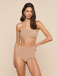 A woman stands against a plain light background with one hand on her hip and a relaxed expression, wearing the Mietta Knit Bandeau Top—a strapless, nude-colored bikini top paired with high-waisted stretch nylon bottoms.