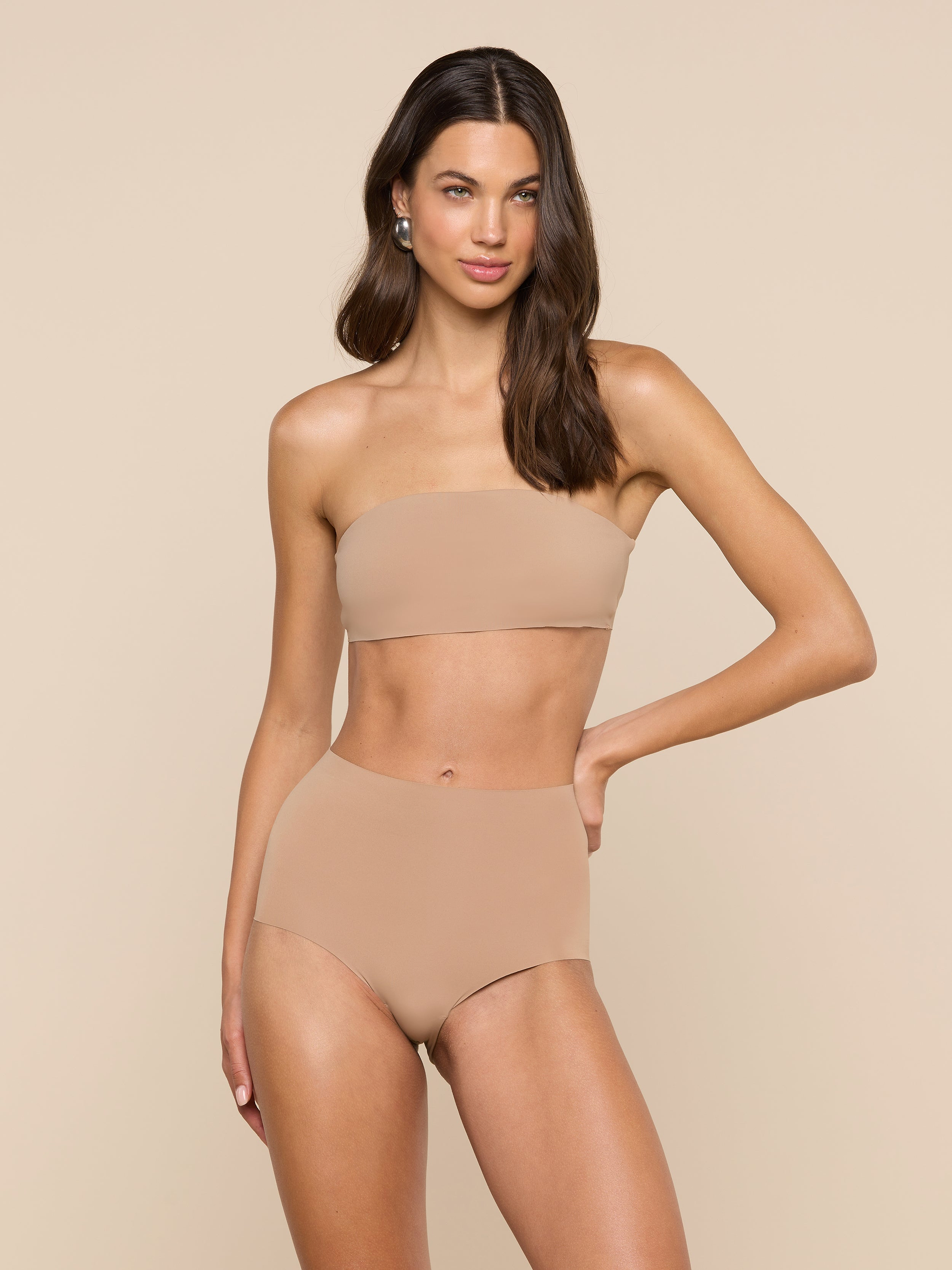A woman stands against a plain light background with one hand on her hip and a relaxed expression, wearing the Mietta Knit Bandeau Top—a strapless, nude-colored bikini top paired with high-waisted stretch nylon bottoms.