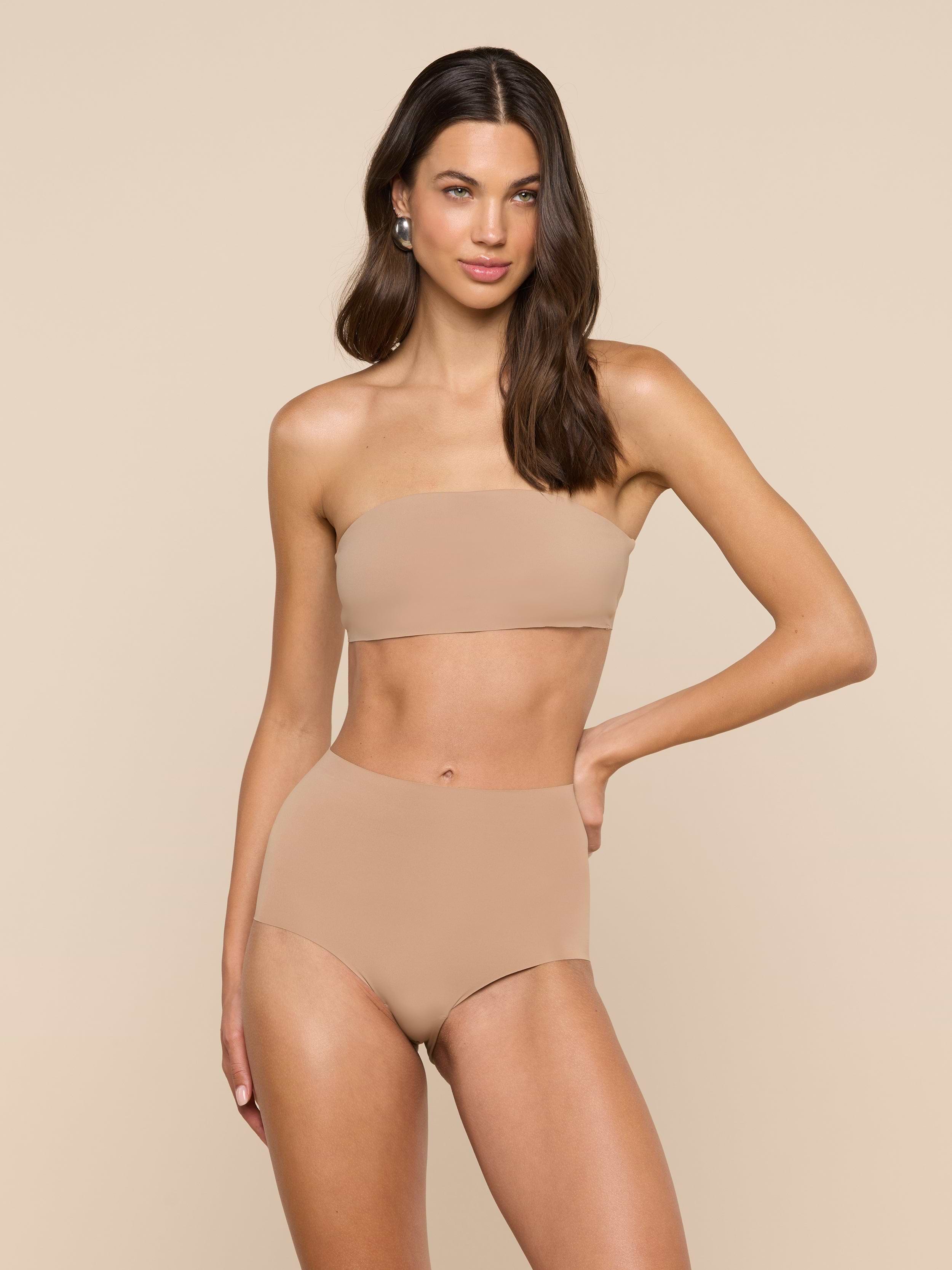 A woman stands against a plain light background with one hand on her hip and a relaxed expression, wearing the Mietta Knit Bandeau Top—a strapless, nude-colored bikini top paired with high-waisted stretch nylon bottoms.