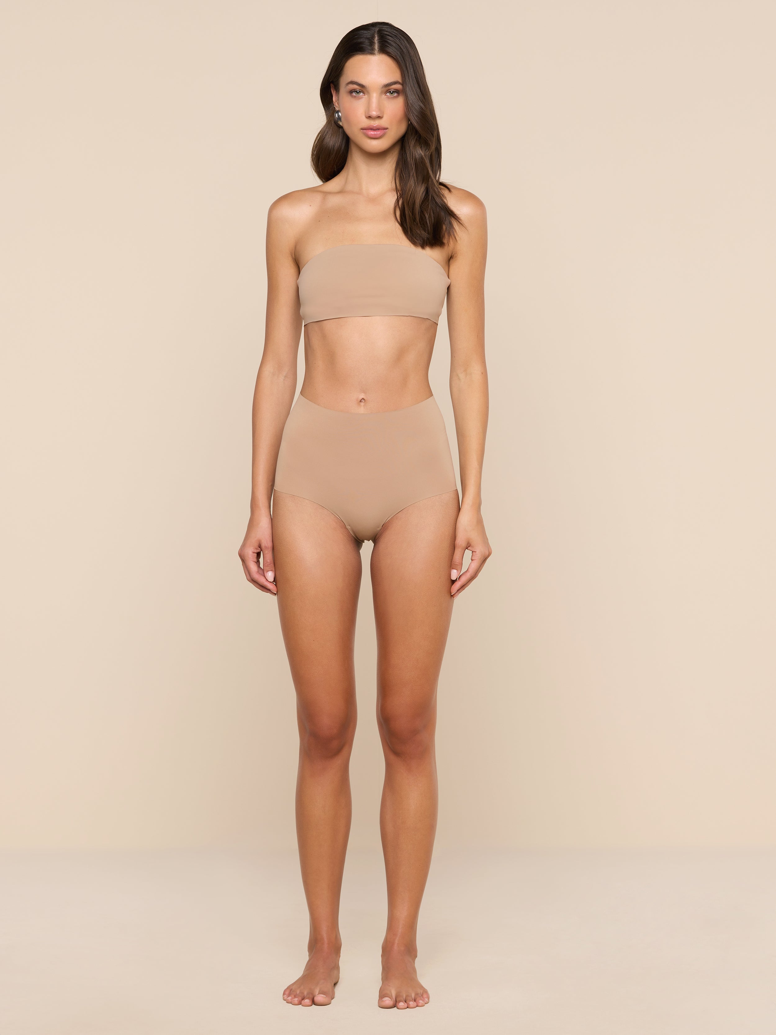 A woman stands barefoot against a plain white background, wearing the Mietta Knit Bandeau Top and matching high-waisted briefs in nude, facing forward with arms relaxed at her sides for a supportive fit.