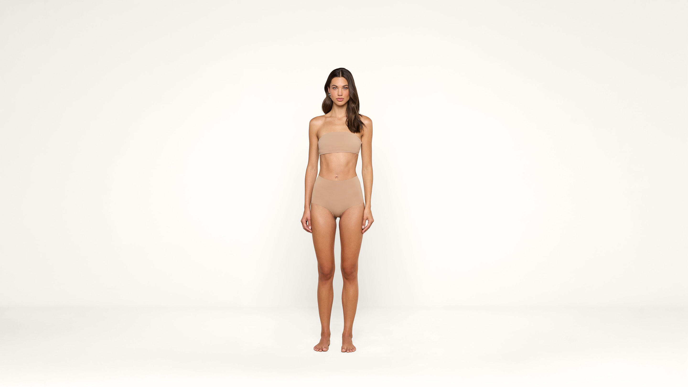 A woman stands against a plain white background, wearing nude-colored undergarments. She faces forward with her arms relaxed at her sides and her expression neutral.