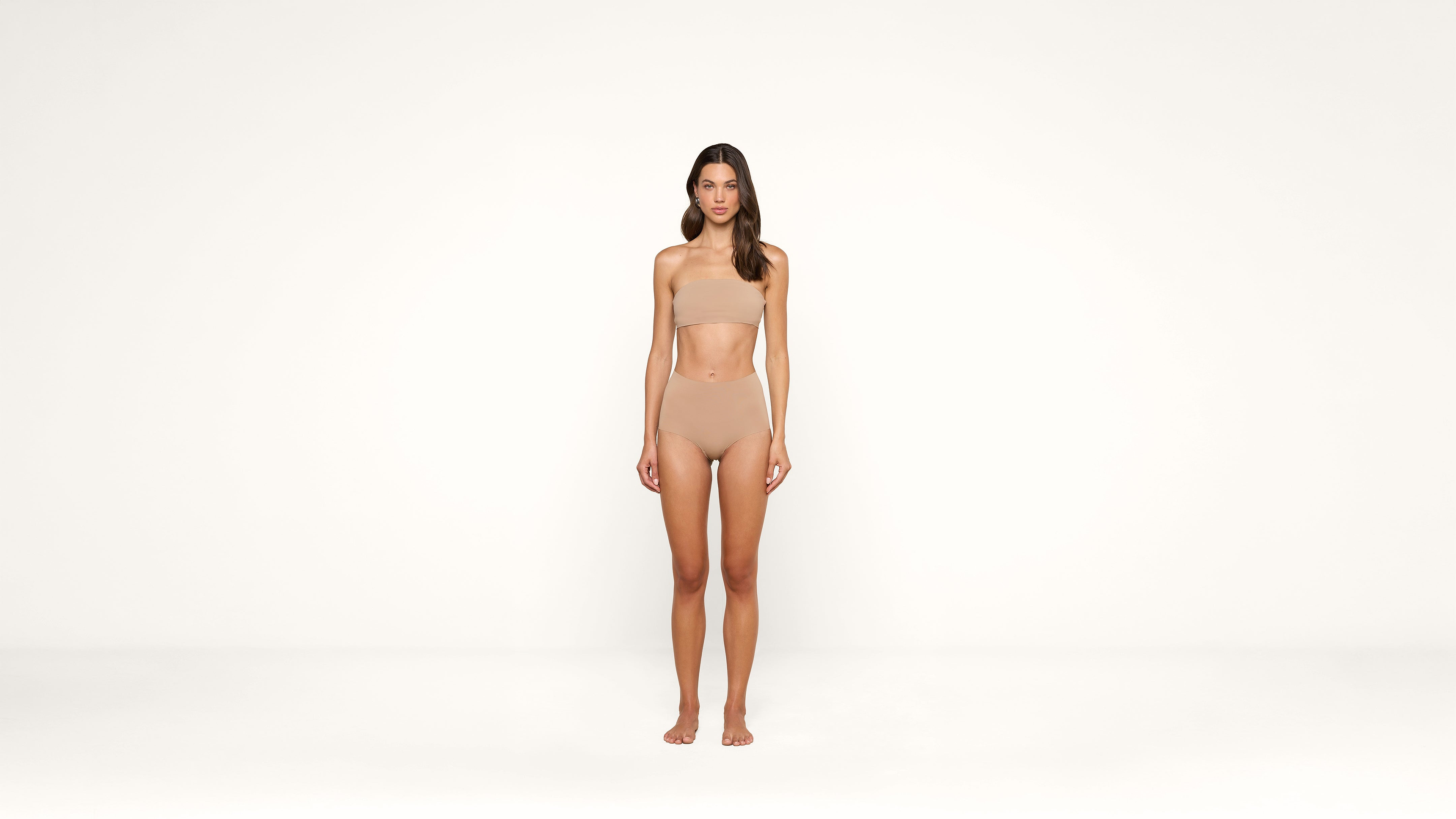 A woman stands against a plain white background, wearing nude-colored undergarments. She faces forward with her arms relaxed at her sides and her expression neutral.