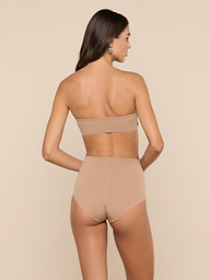 A woman stands facing away, wearing the Mietta Knit Bandeau Top in a strapless beige design, paired with high-waisted underwear. The set is crafted from stretch nylon for a supportive fit, set against a plain, light-colored background.