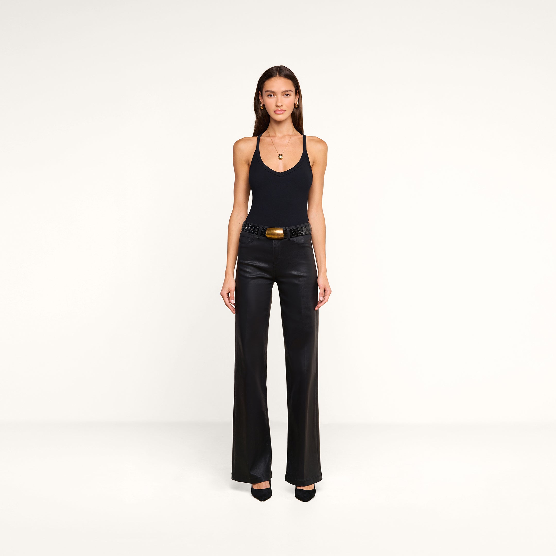 A woman stands against a plain white background, wearing the Wrenna Racerback Bodysuit in black rib fabric, paired with black leather pants, a black belt with a large buckle, and black heels. Her long straight hair frames her neutral expression.
