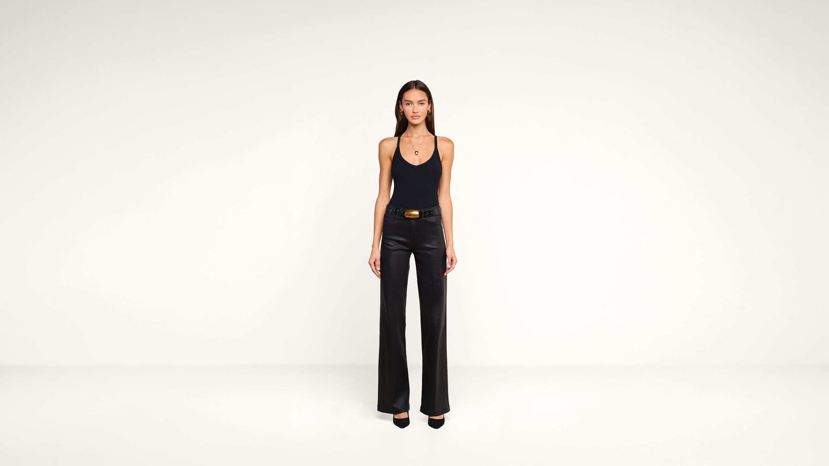 A woman stands facing forward in a minimal studio, wearing a black tank top, black wide-leg pants, a belt, and black heels. The background is plain white.