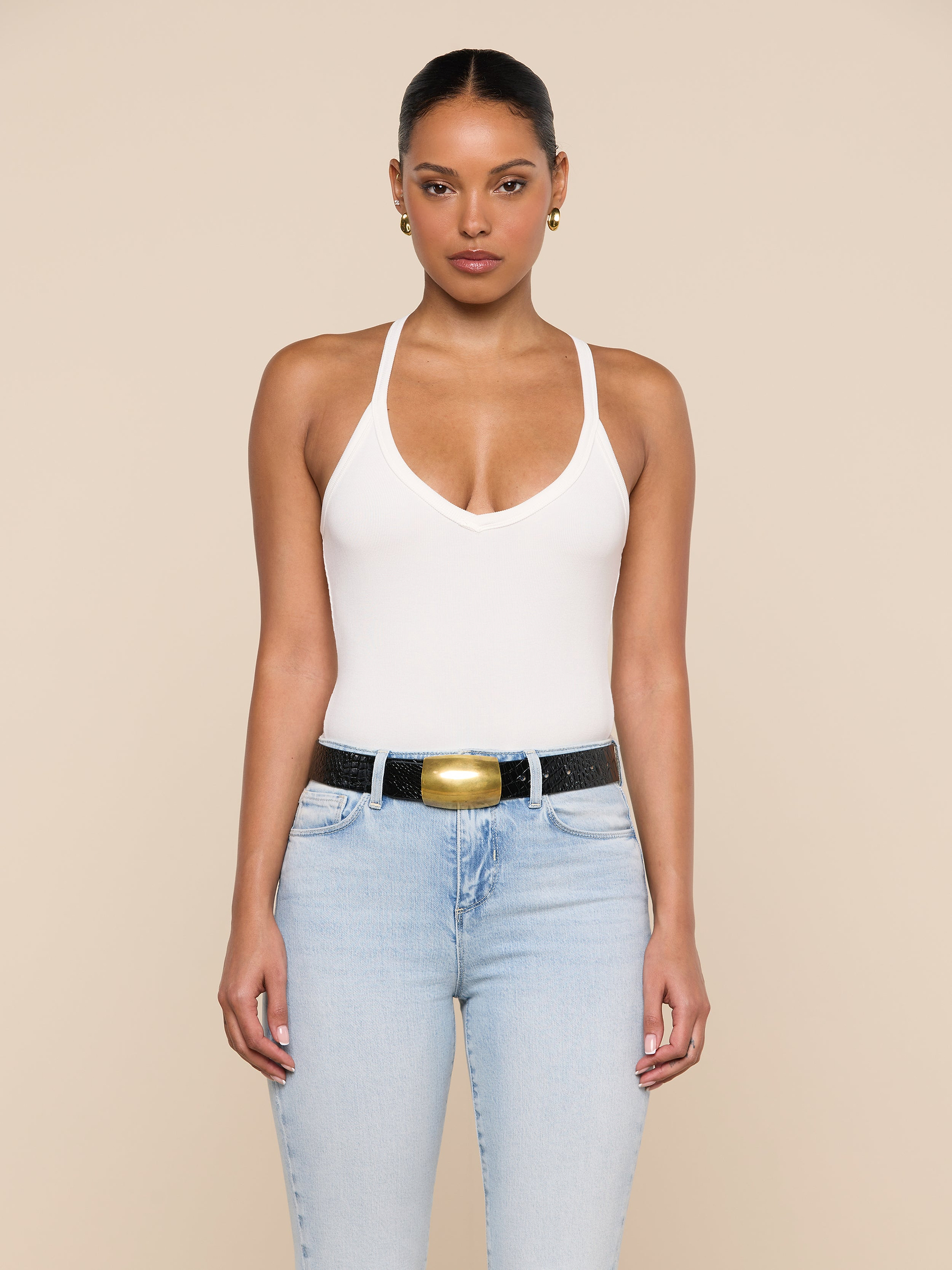 A woman stands facing forward against a plain light background, wearing the Wrenna Racerback Bodysuit in white rib fabric, paired with light blue jeans and a black belt with a large gold buckle.