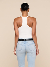A person with hair in a bun, wearing the Wrenna Racerback Bodysuit in white rib fabric, paired with light blue jeans and a black belt, stands with their back to the camera against a plain, light background.