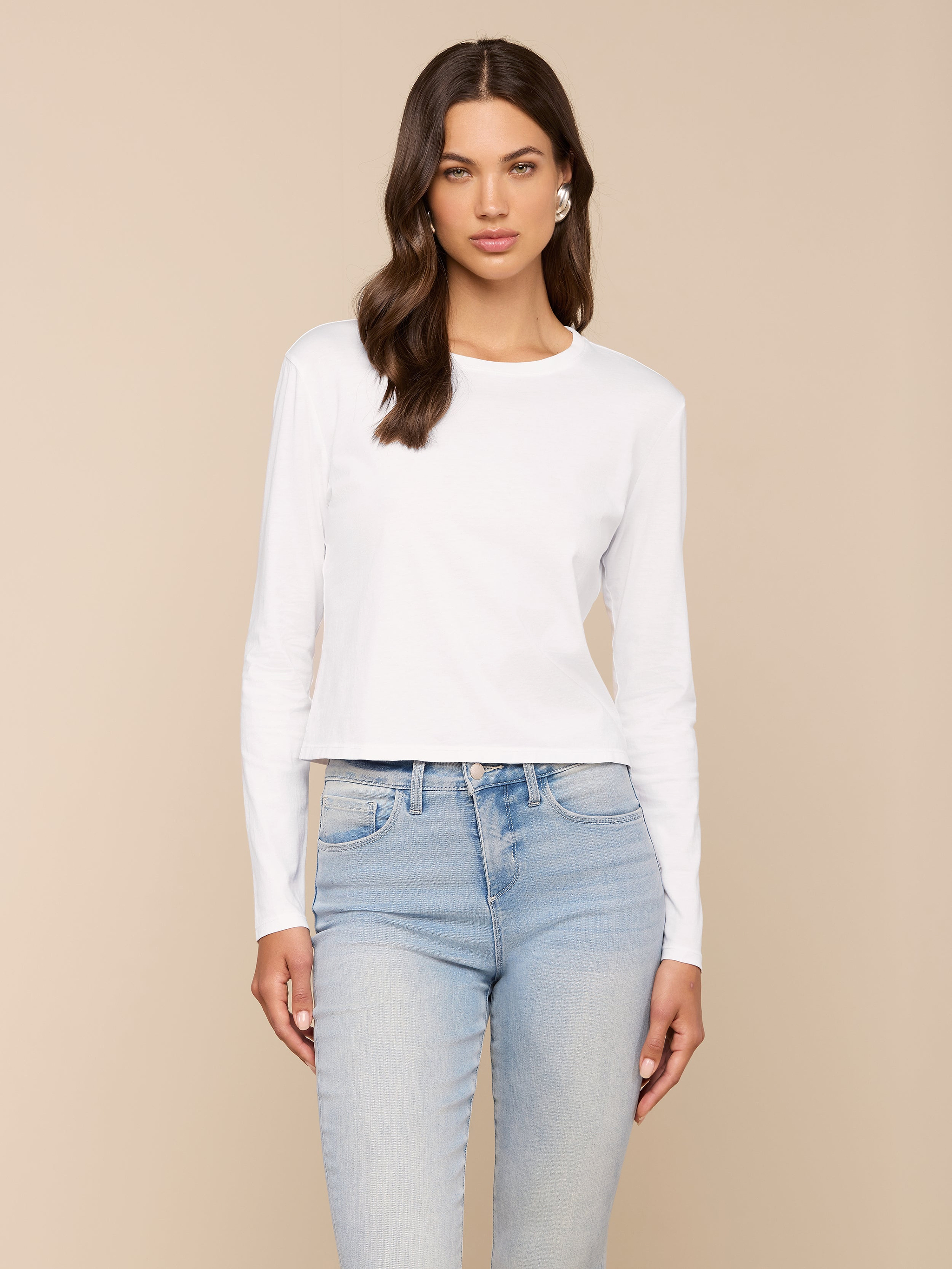 A woman with long brown hair wears the Rojene Cotton Tee, a relaxed fit plain white long sleeve shirt, paired with light blue jeans and standing against a plain light background.