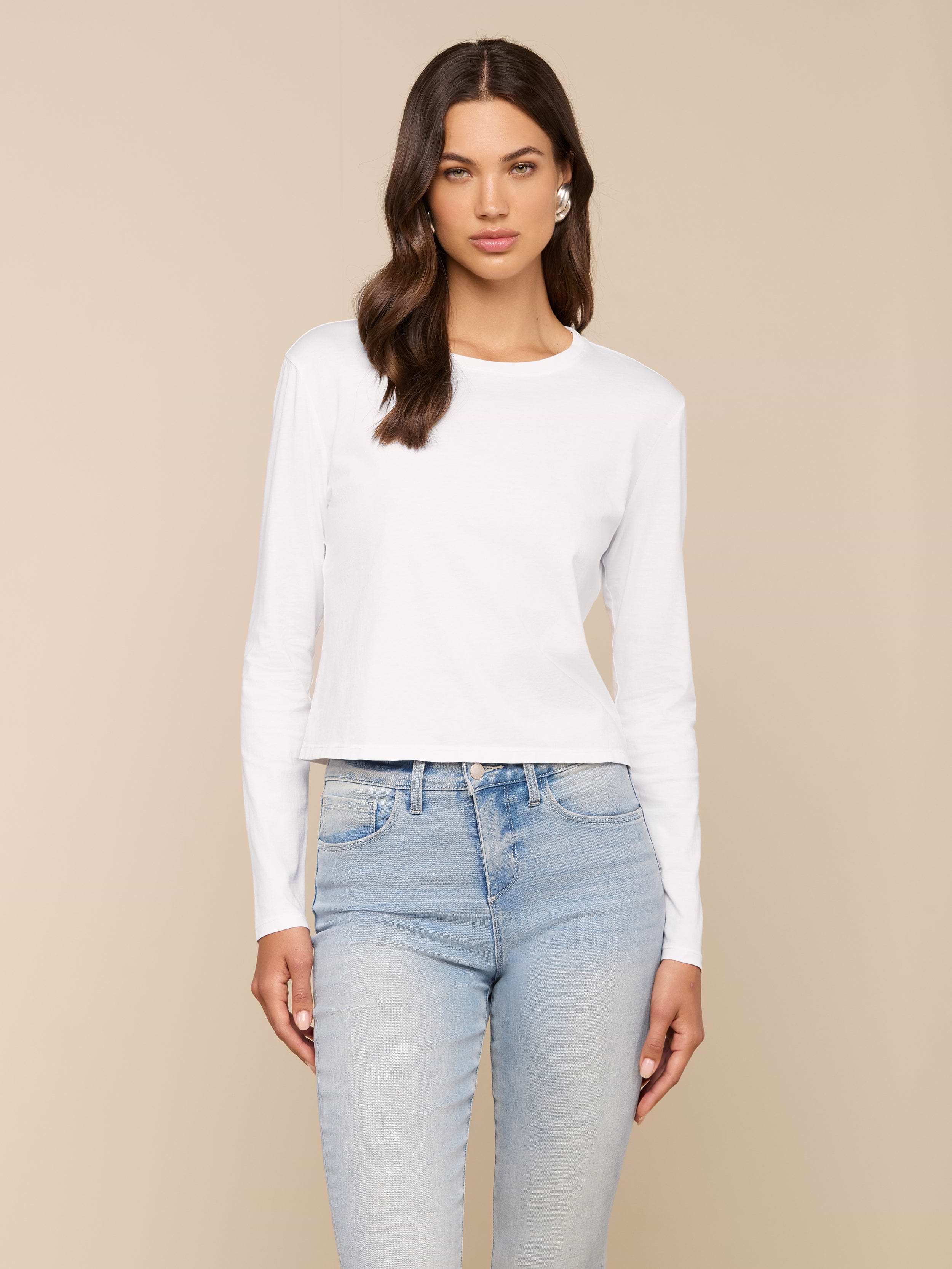 A woman with long brown hair wears the Rojene Cotton Tee, a relaxed fit plain white long sleeve shirt, paired with light blue jeans and standing against a plain light background.