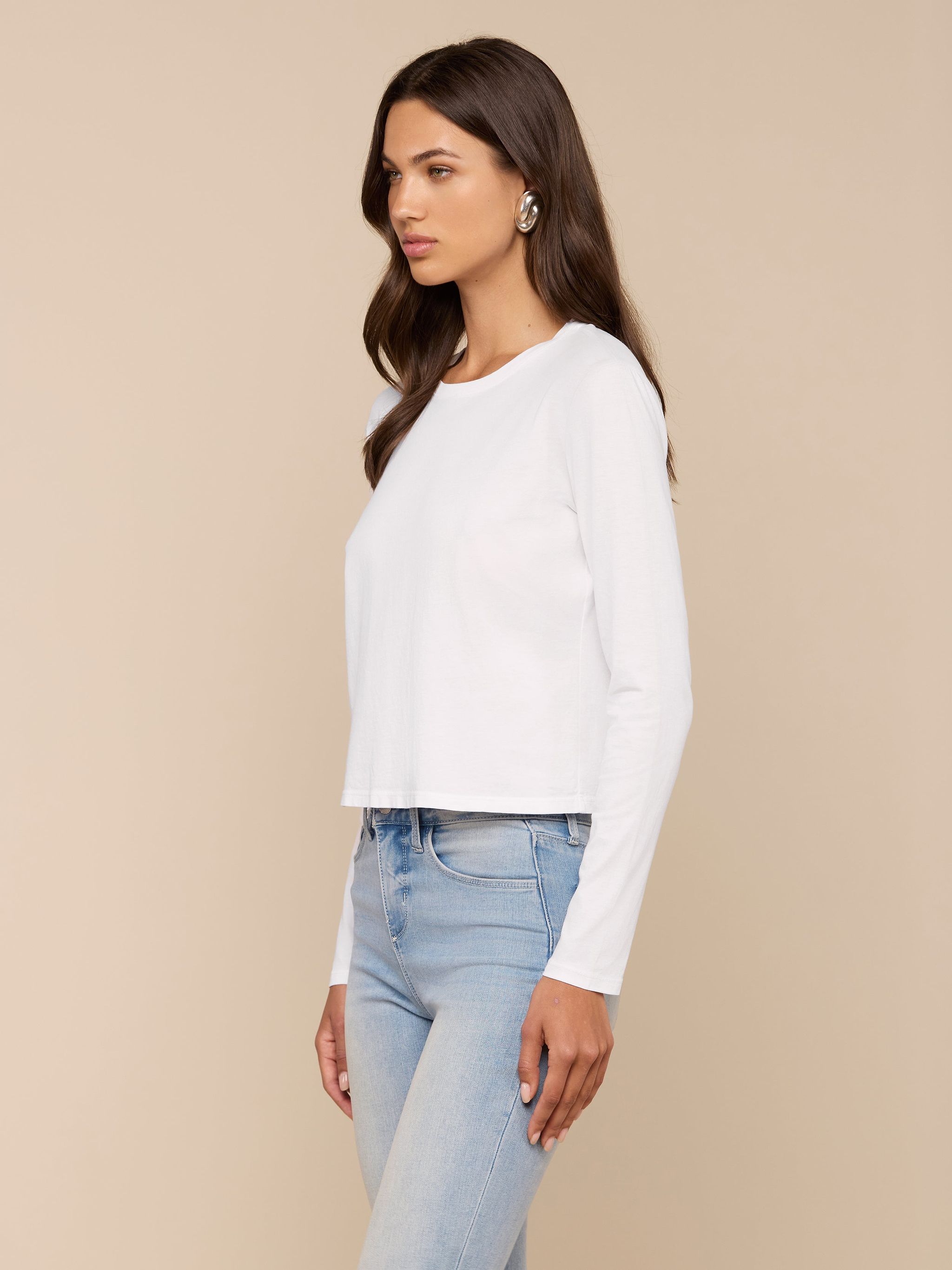 A woman with long brown hair wearing a relaxed fit, white Rojene Cotton Tee and light blue jeans stands facing sideways against a plain, light background.