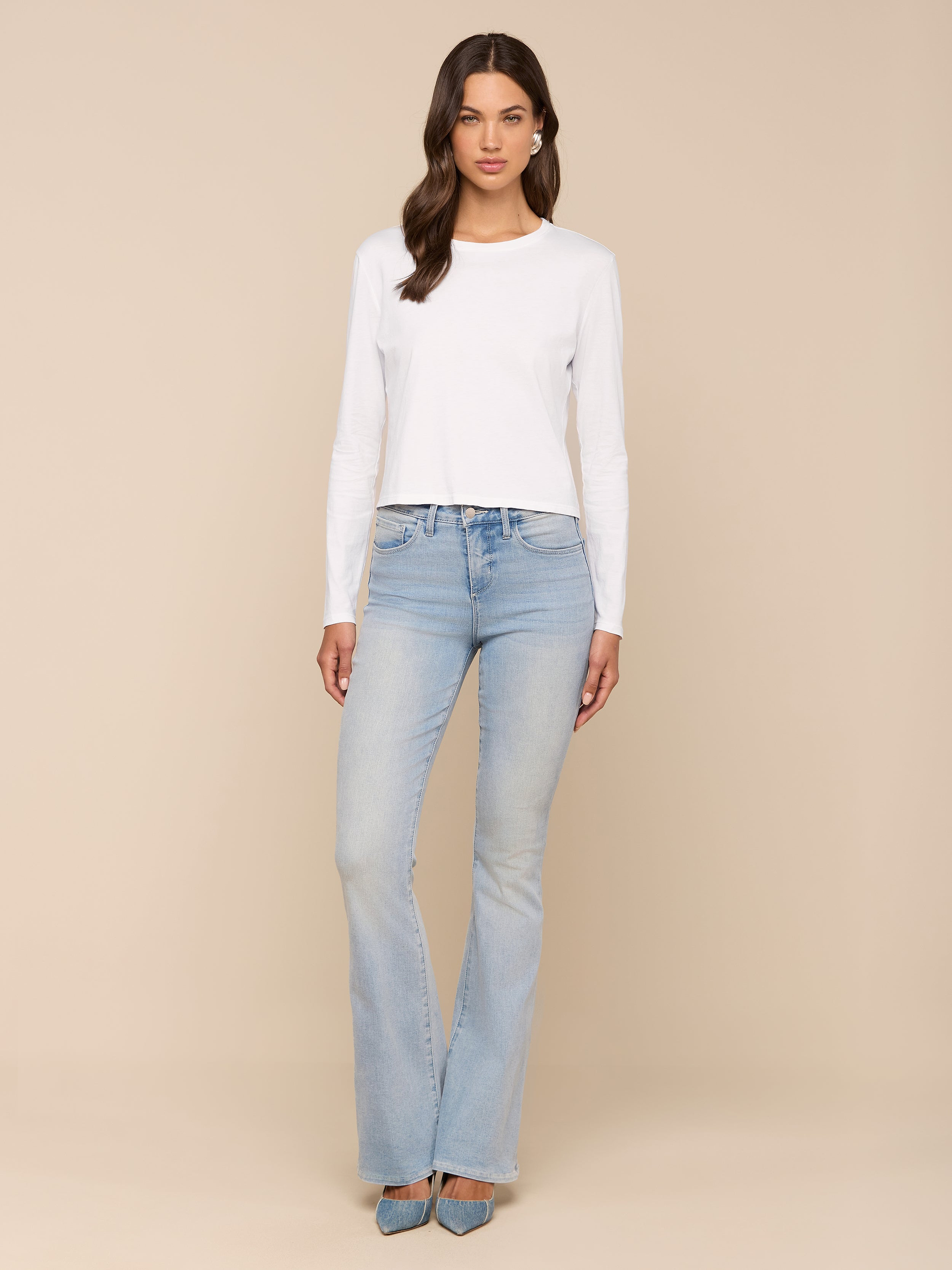 A woman with wavy brown hair and a neutral expression stands against a plain background, wearing the relaxed fit Rojene Cotton Tee paired with light blue flared jeans and pointed-toe shoes.