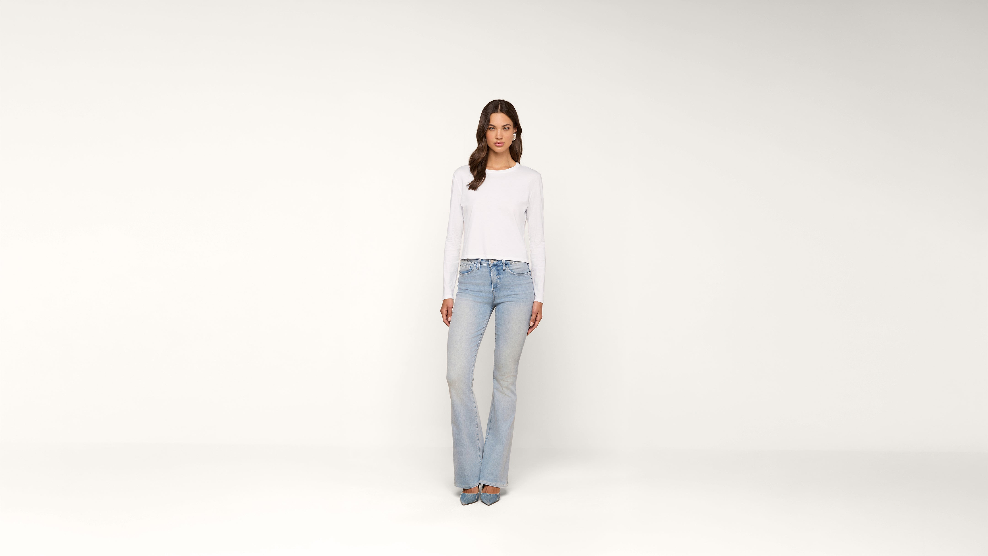 A woman stands against a plain white background, wearing a white long-sleeve shirt, light blue jeans, and gray heels. She faces forward with a relaxed expression and hands by her sides.